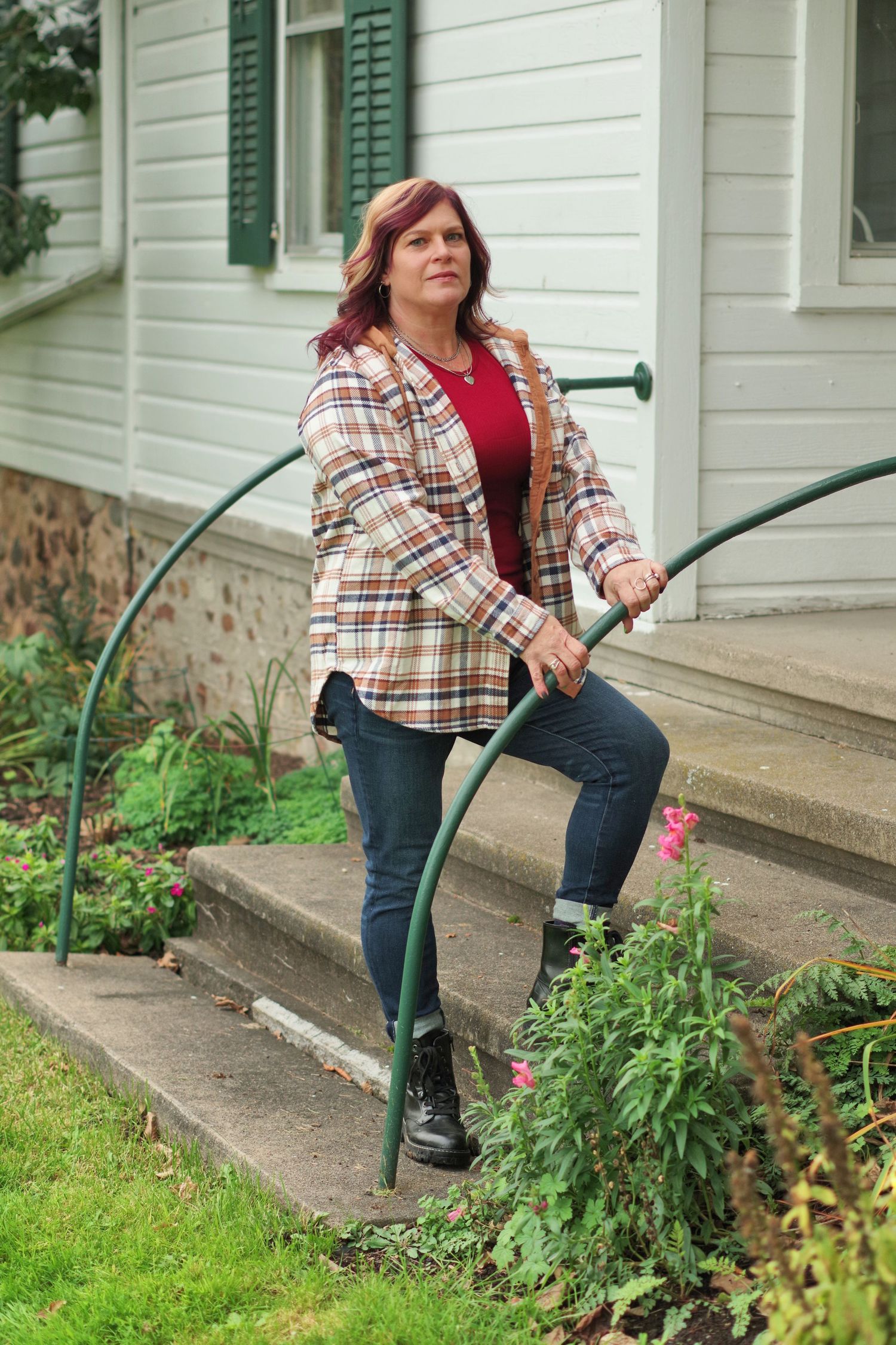 Person in plaid shirt and boots poses on house steps with green handrail.