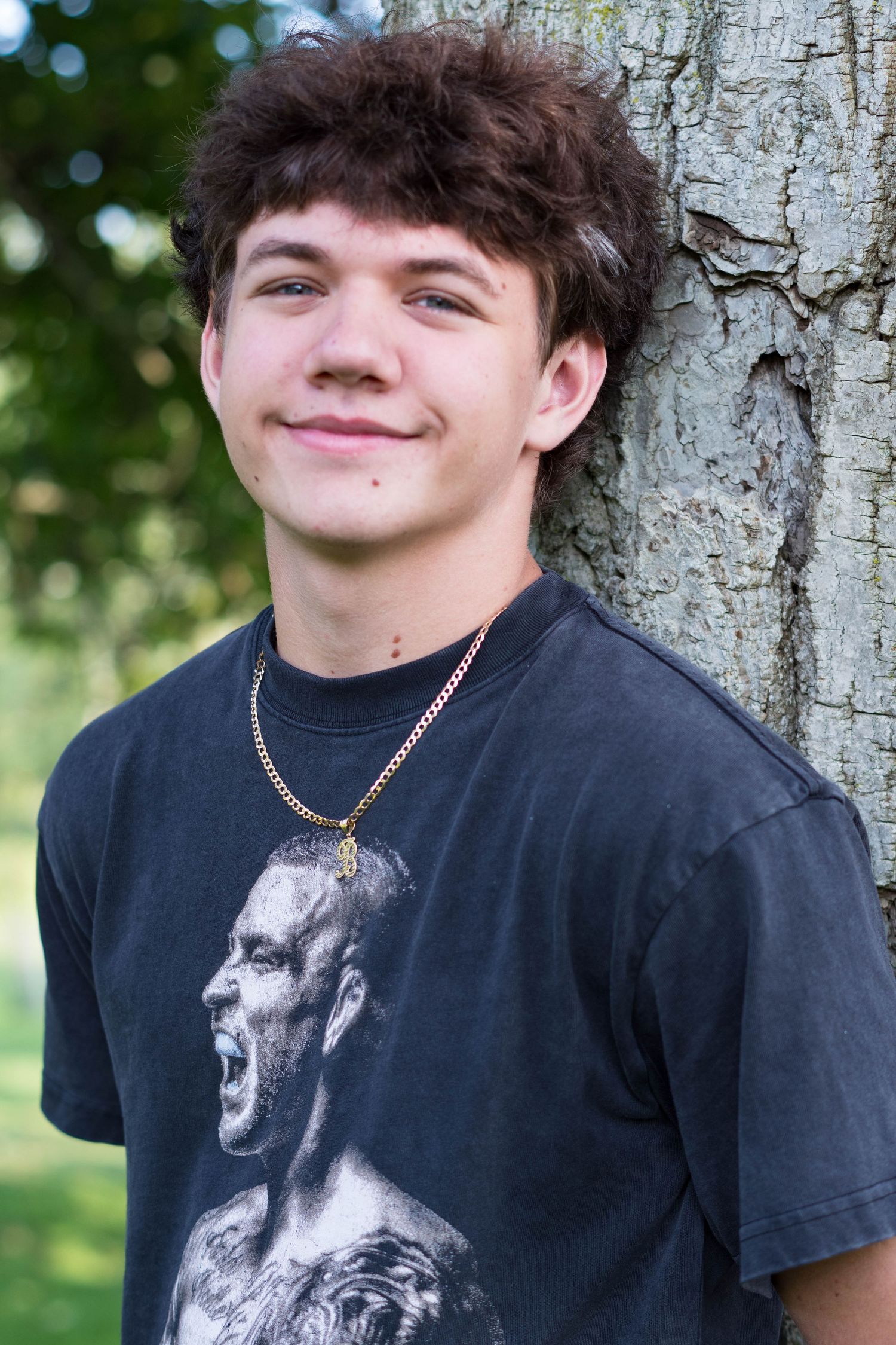 Young man with curly hair leaning against a tree, wearing a graphic tee and gold chain necklace outdoors.