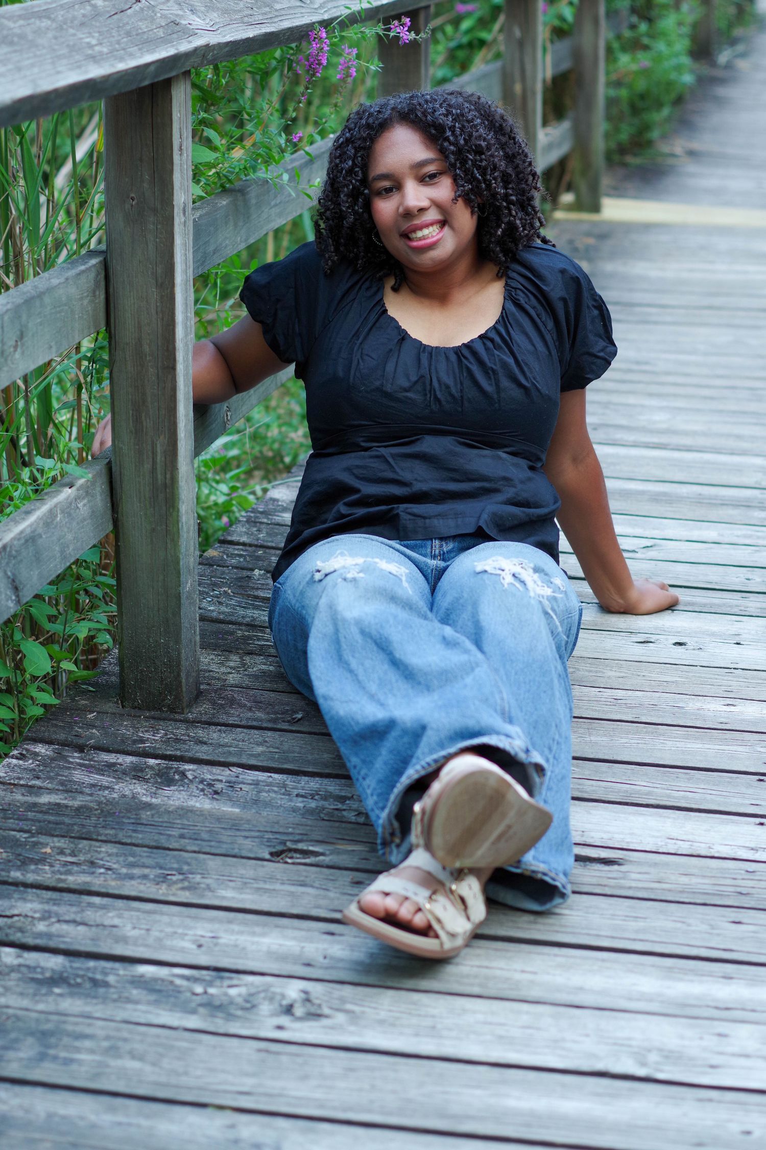 Person relaxing on a wooden boardwalk surrounded by greenery wearing jeans and a black top.