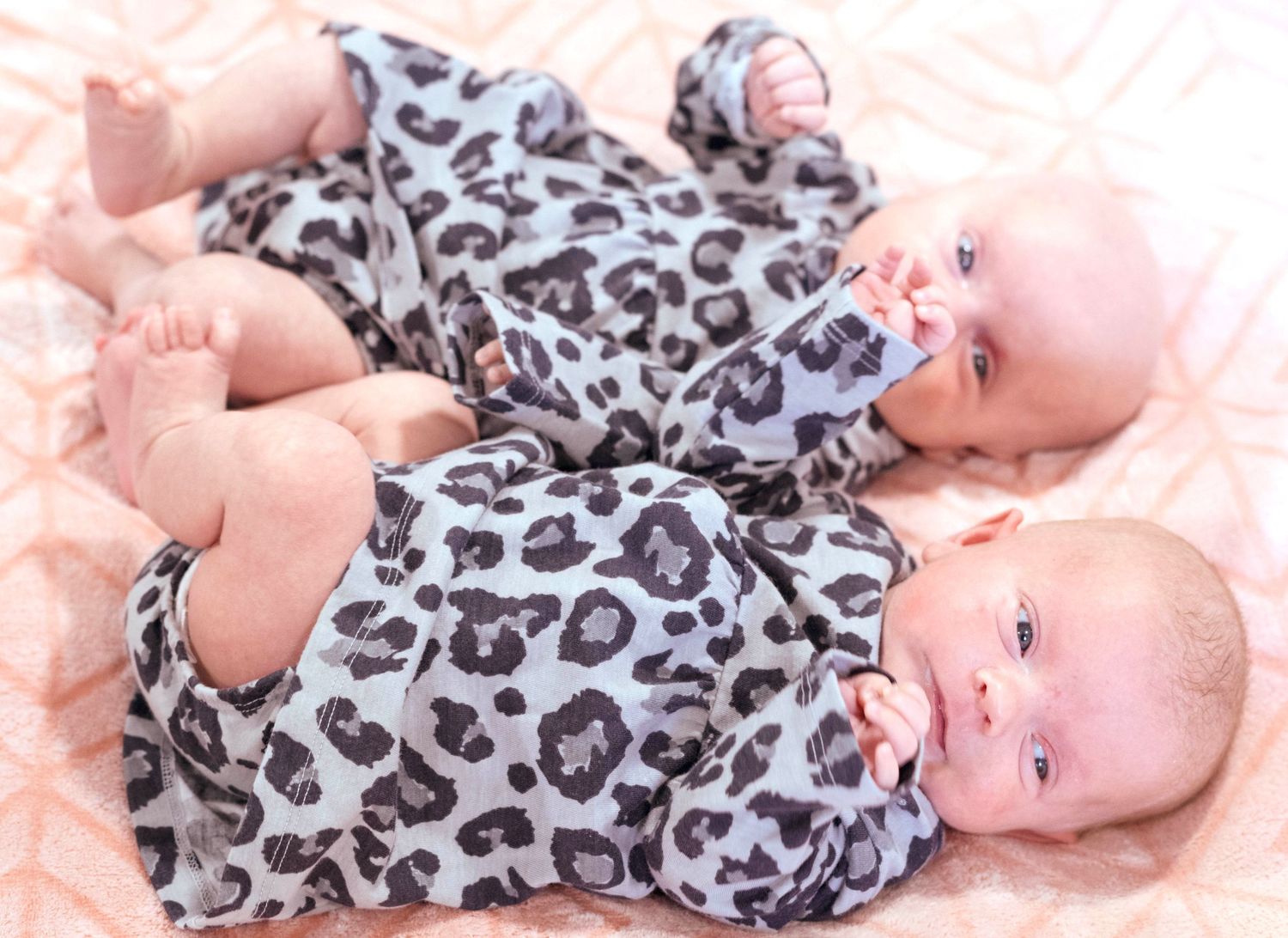 Two babies wearing matching leopard print outfits lying on pink bedding.