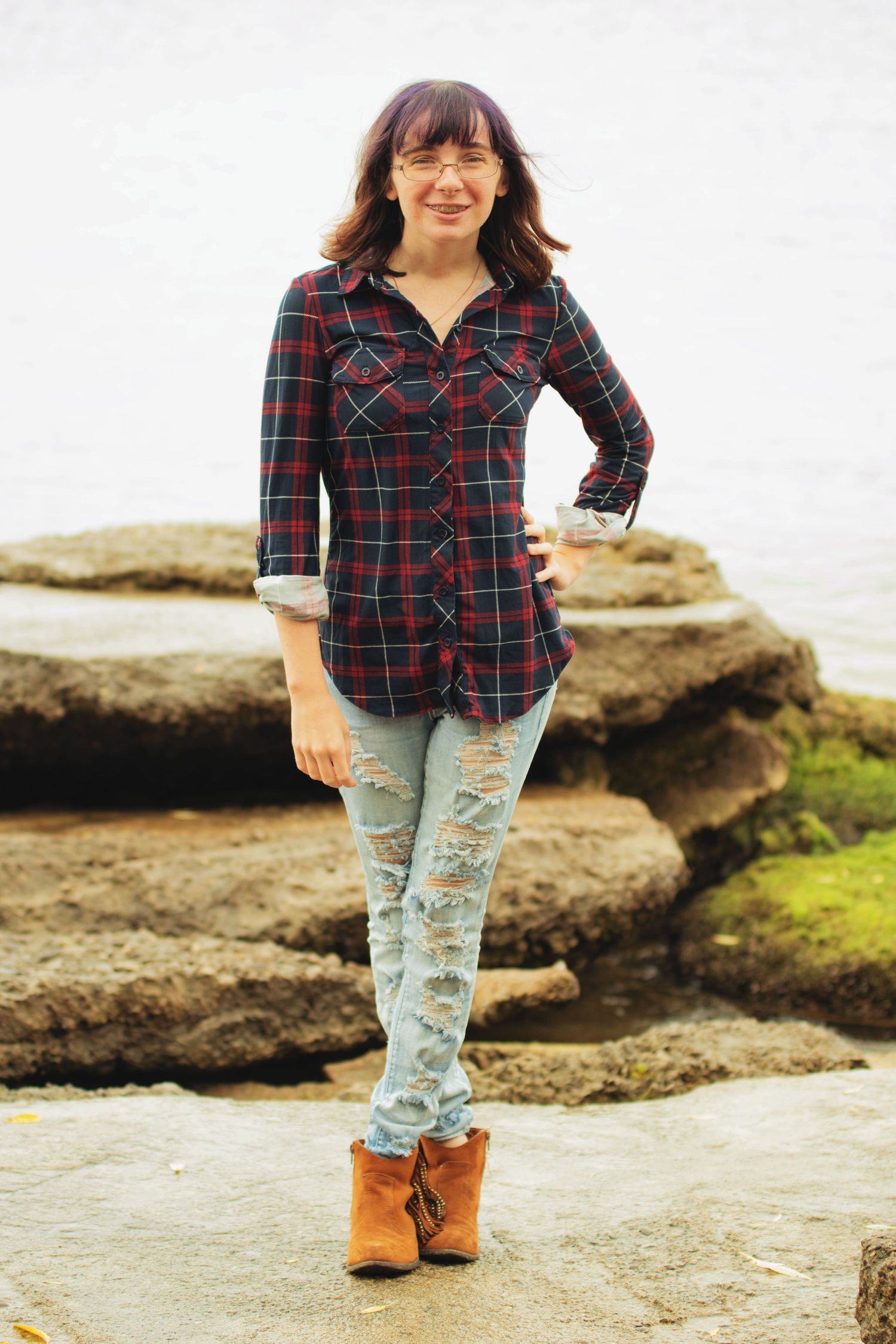 Person wearing plaid flannel shirt, distressed jeans and brown boots poses on coastal rocks.