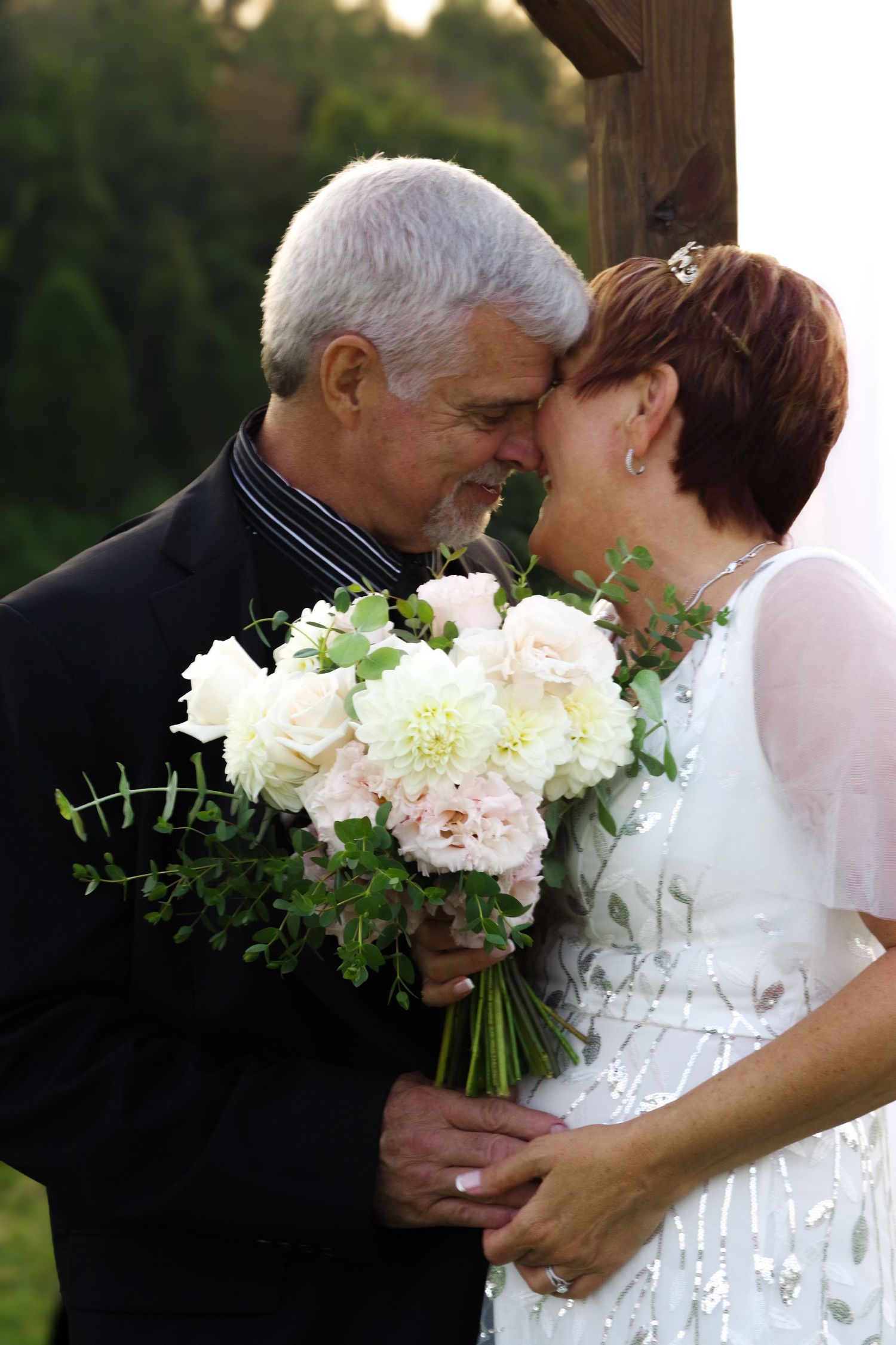 A romantic wedding moment with a couple sharing a tender moment and holding a white floral bouquet.