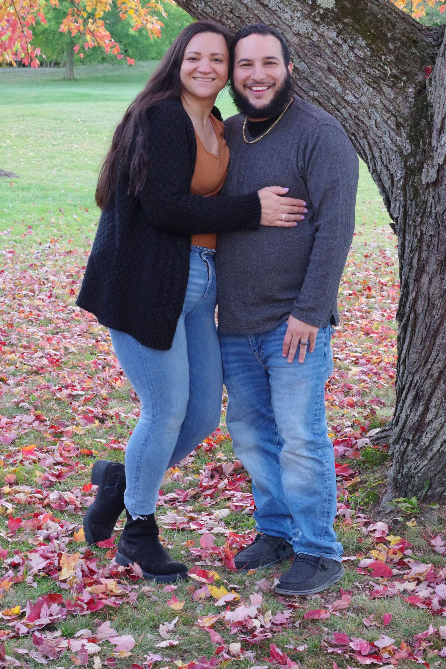 Couple poses together by tree trunk surrounded by colorful autumn leaves on ground.