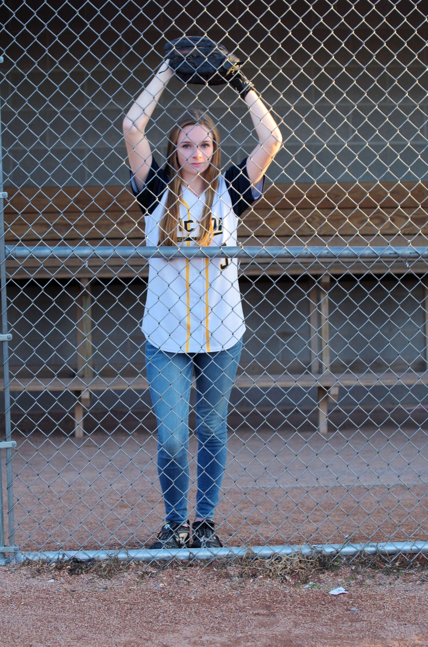 Baseball player in white jersey and jeans poses with glove by chain link fence on field.