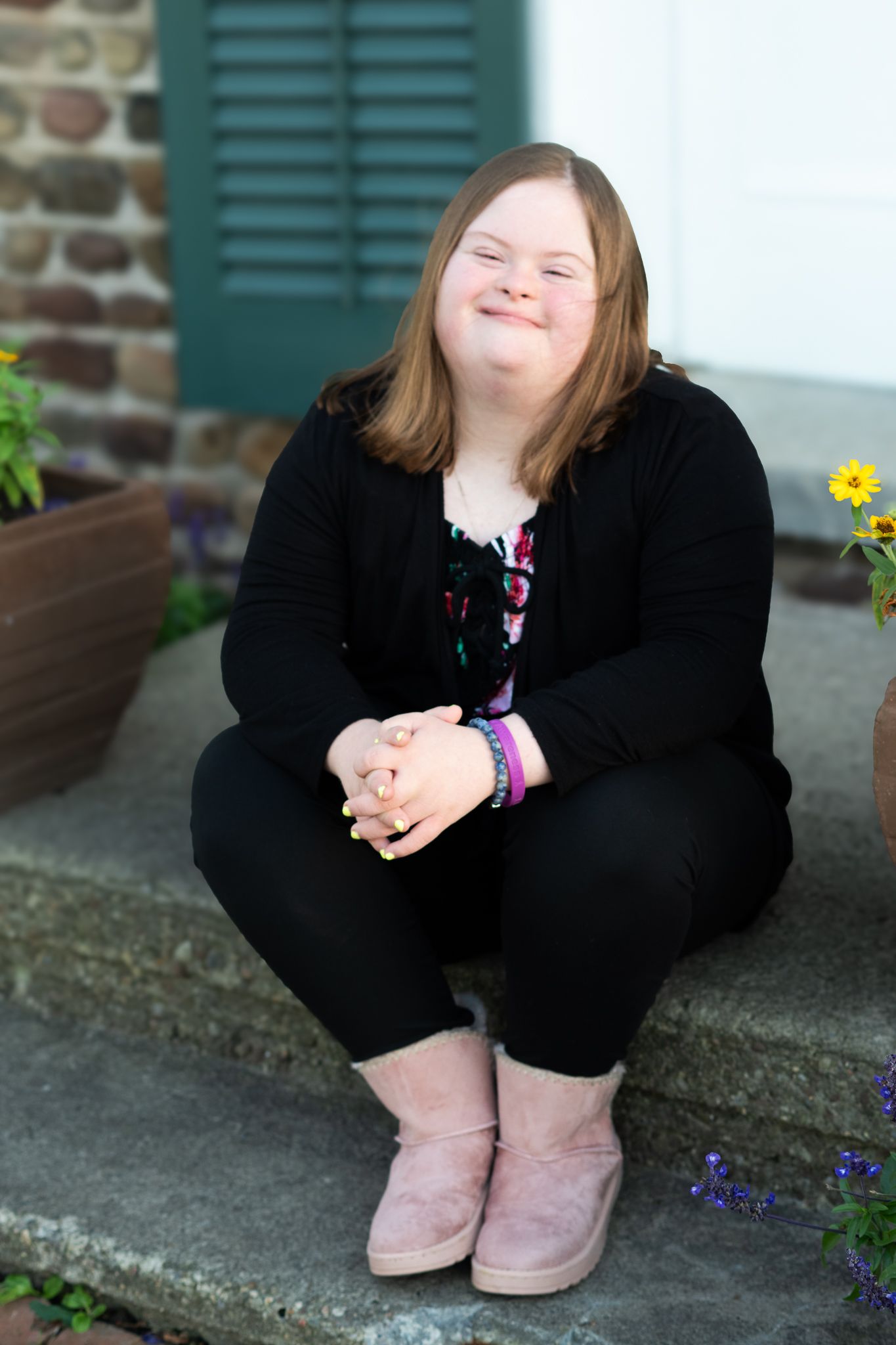 Smiling teenage girl with Down syndrome sitting on stone steps outdoors in black outfit and pink boots.