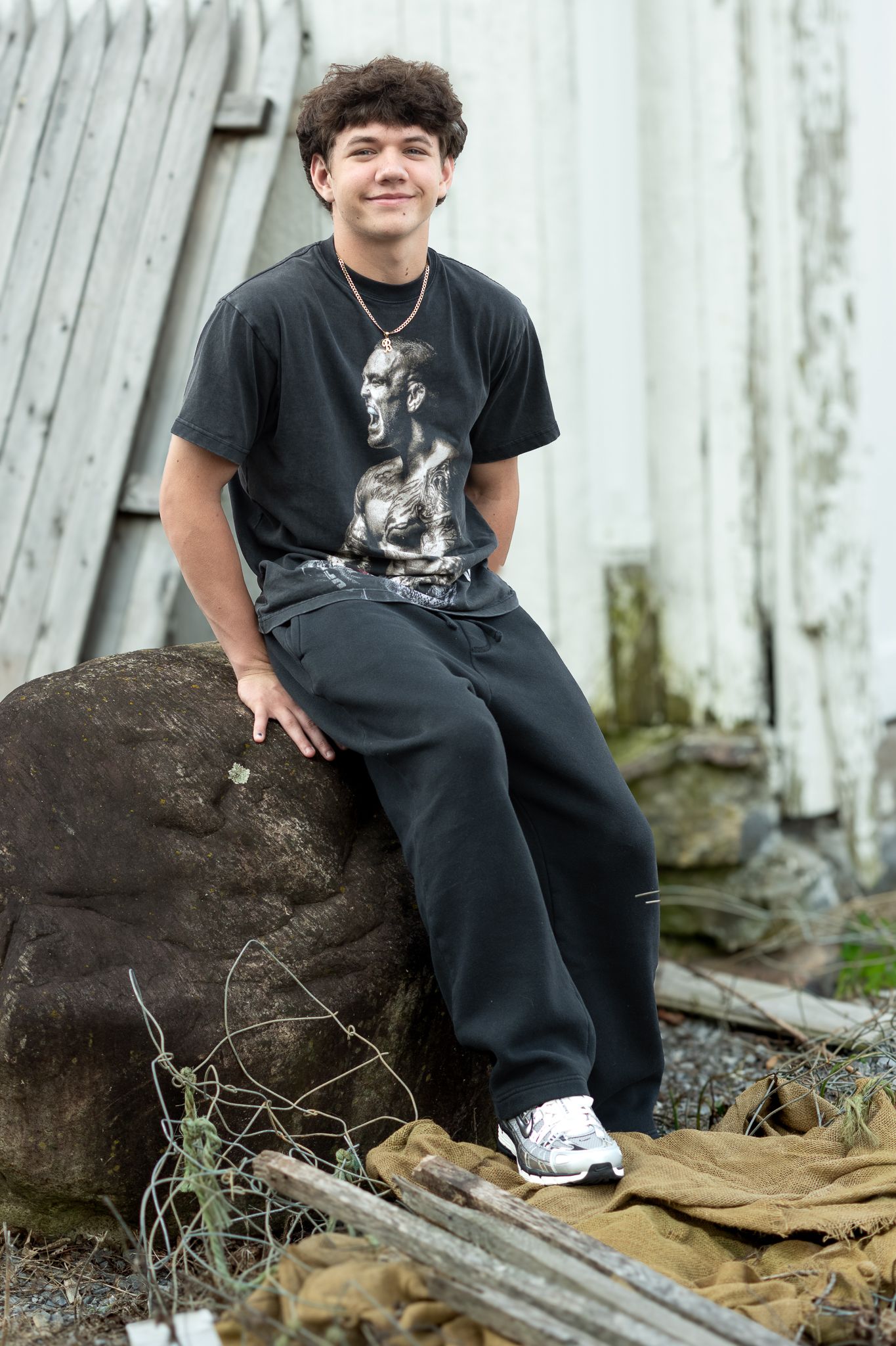 A smiling teenage boy in black outfit sits on a rock near a rustic barn wall outdoors.