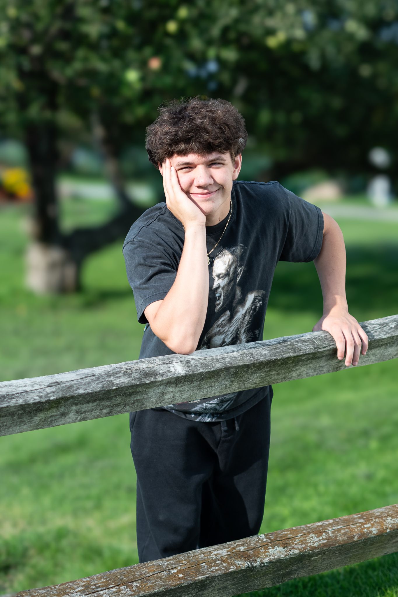 A teenage boy in a black graphic tee leans on a wooden fence, smiling in a lush green park.