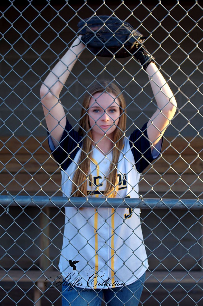 Young female softball player holding glove above head behind chain-link fence in team jersey.