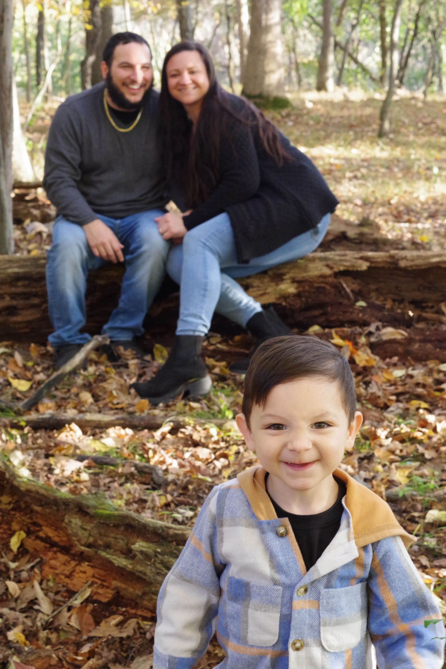 Family sitting on fallen log in wooded area with autumn leaves on ground.