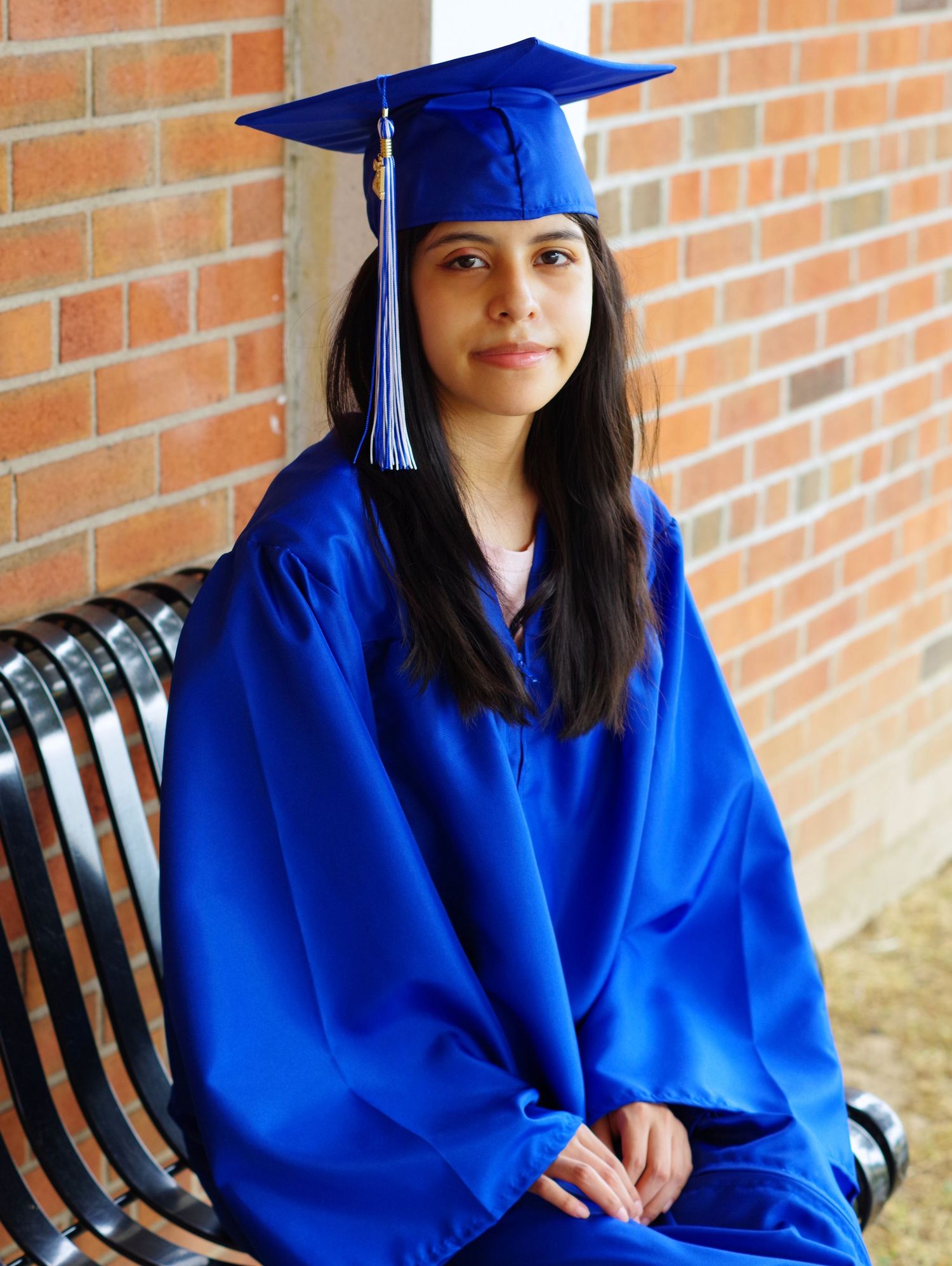 Graduate in traditional blue cap and gown sitting on bench near brick building.