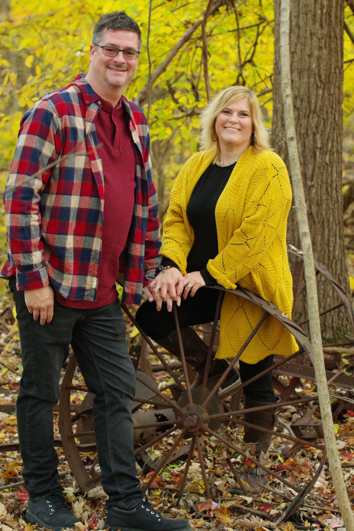 Couple in plaid and yellow sweater posing with vintage farm equipment in fall foliage.