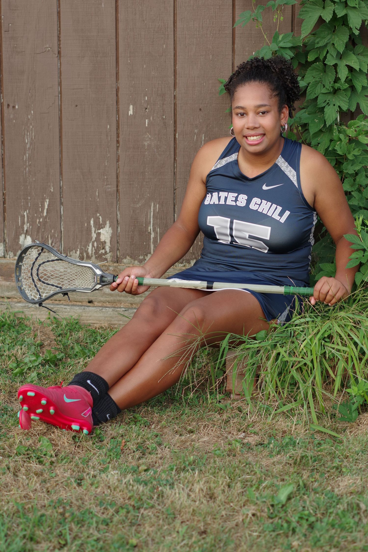 Athlete in navy Gates Chili jersey sits on grass holding a lacrosse stick near a brown fence.