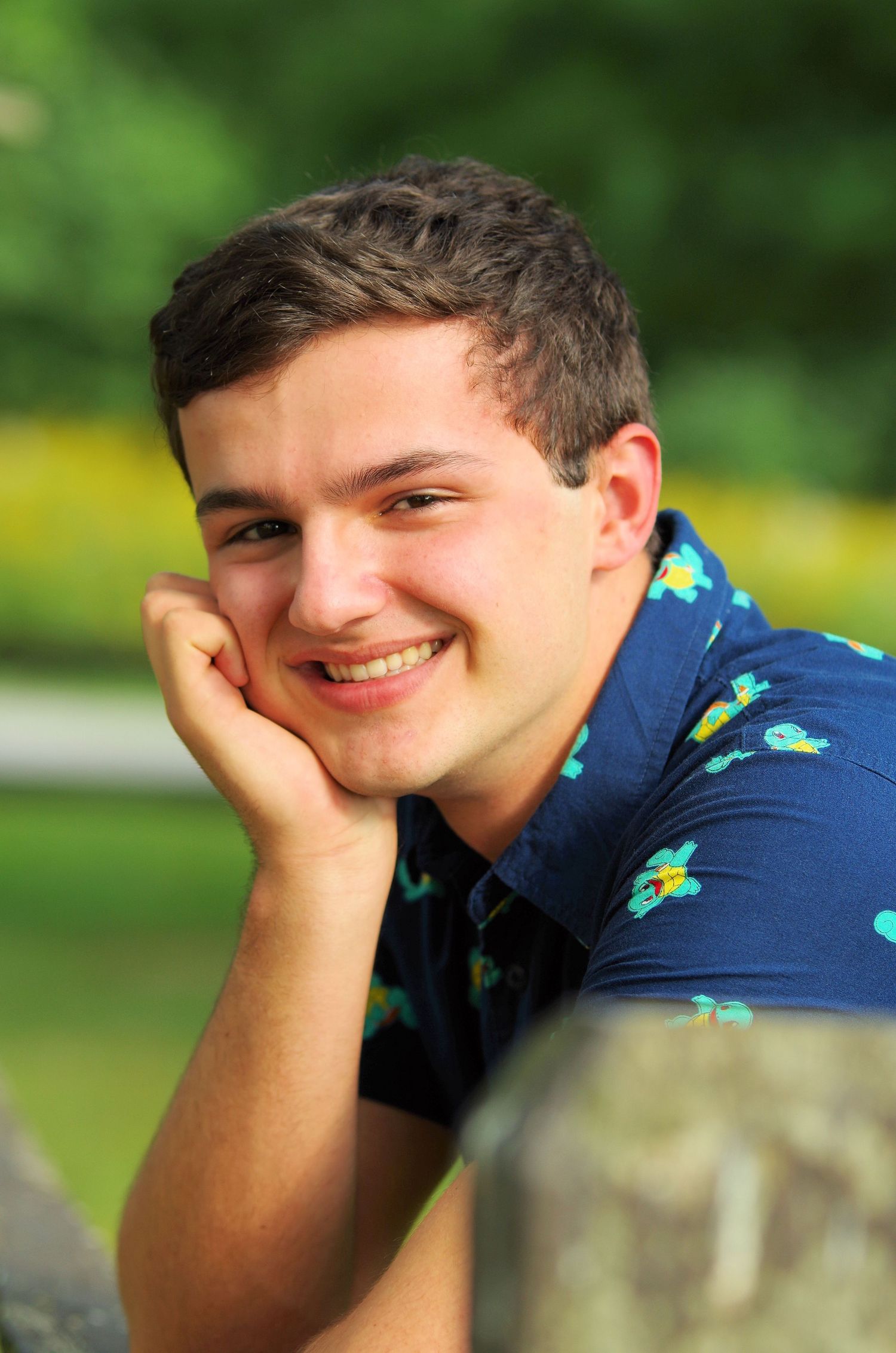 Relaxed outdoor portrait poses with hand resting on chin against green natural backdrop.
