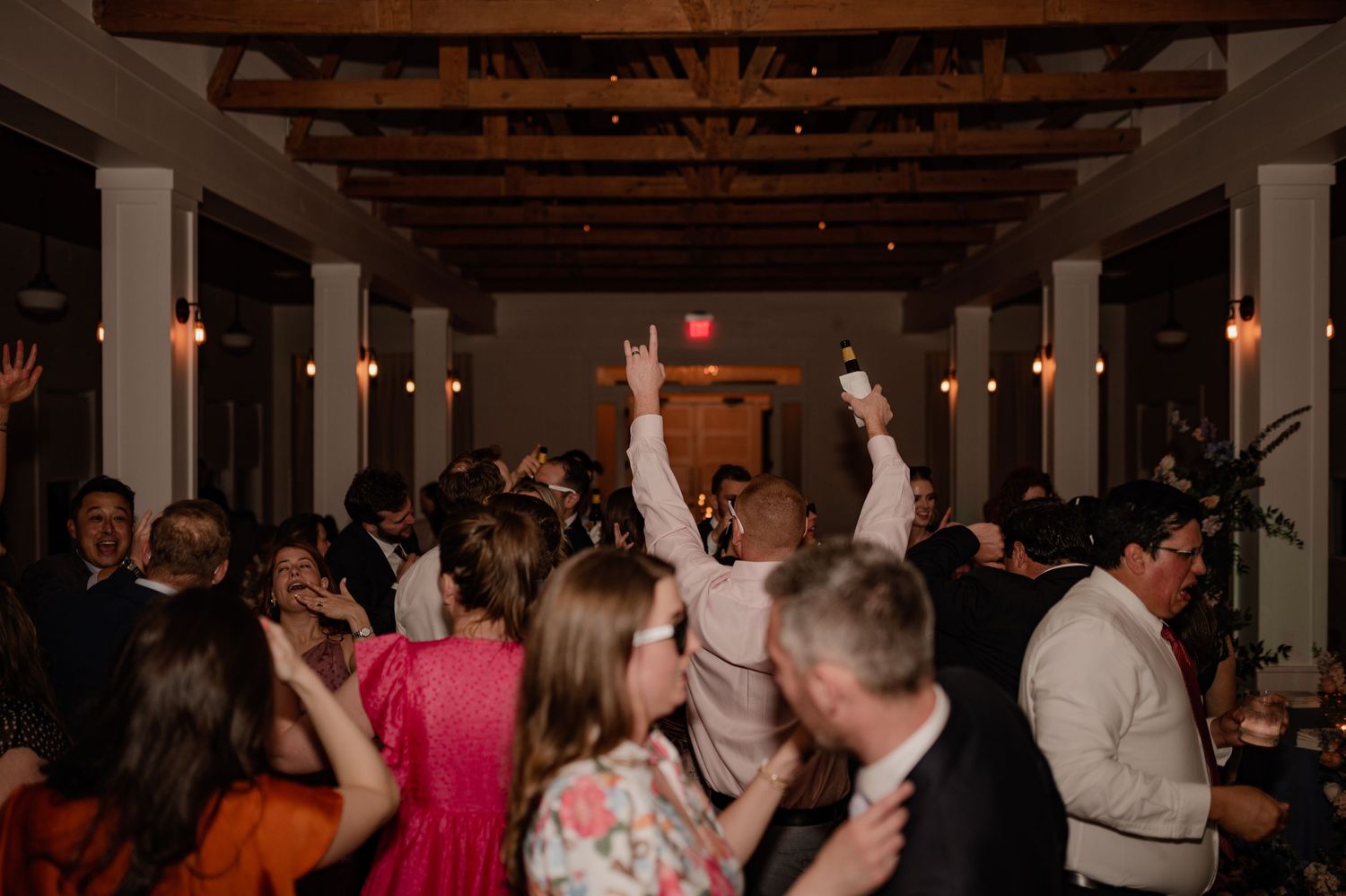 Wedding reception guests dance and celebrate on a crowded dance floor under wooden beams and mood lighting.