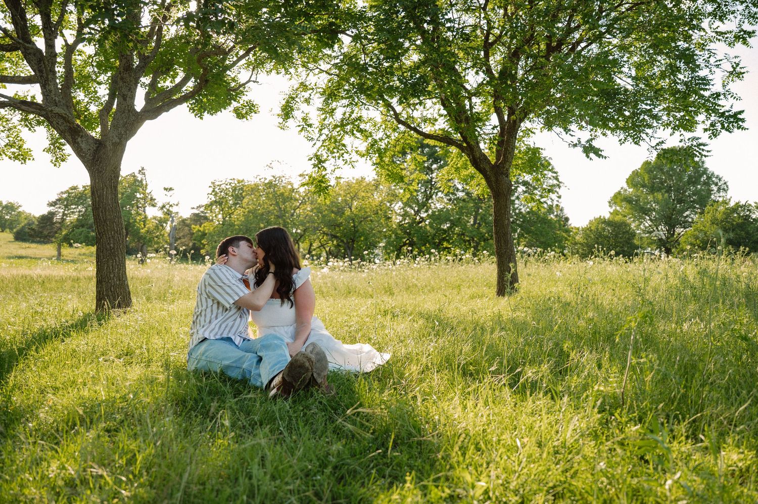 Couple sitting in a field of wildflowers with the lake and sunset behind them for an engagement session in East Dallas, Texas