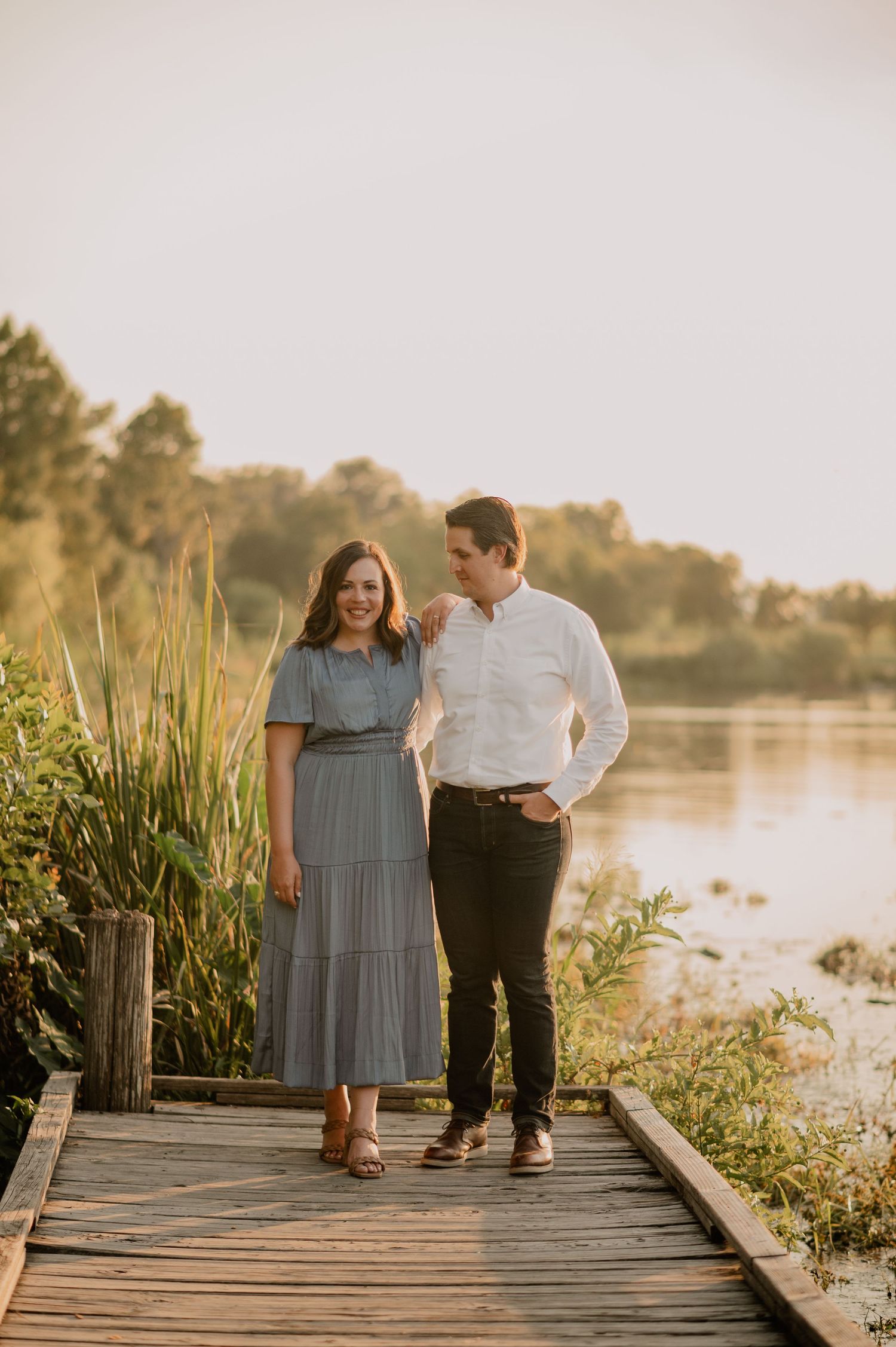 Couple standing on a wooden dock at White Rock Lake at golden hour, woman in blue dress, man in white shirt.
