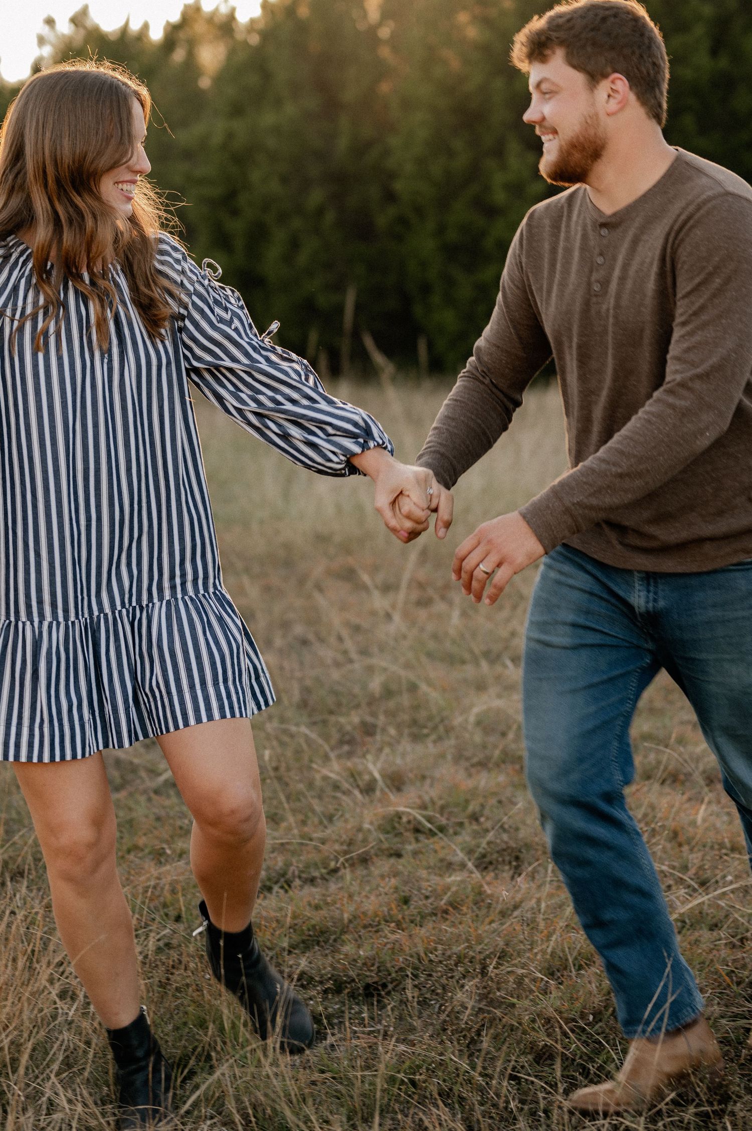 Married couple joyfully running and holding hands during a casual fall couple session in a large tree lined field in during golden hour
