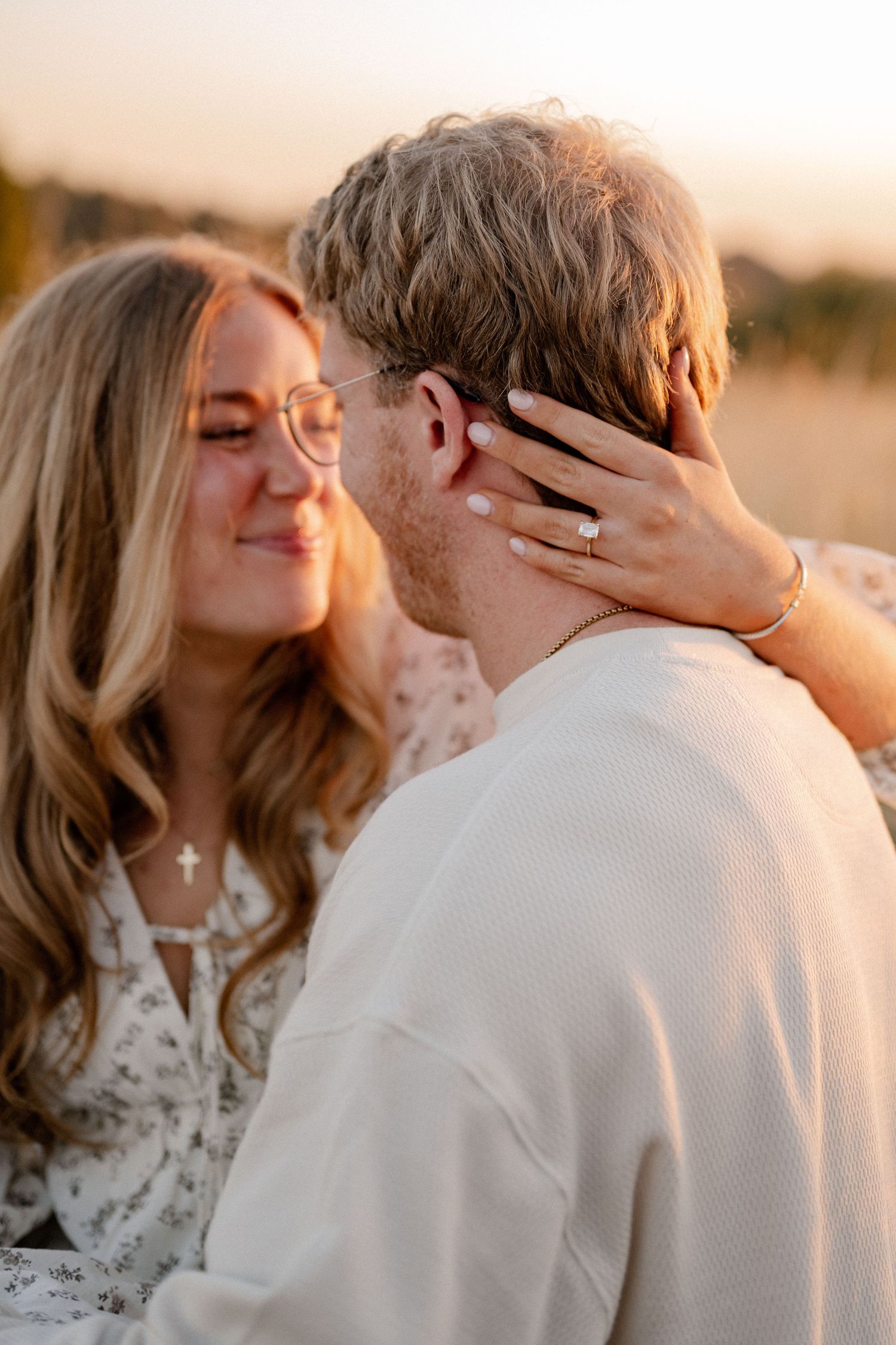 Candid shot of an engaged couple, with a focus on the engagement ring, embracing while sitting in a large field overlooking a lake in Dallas