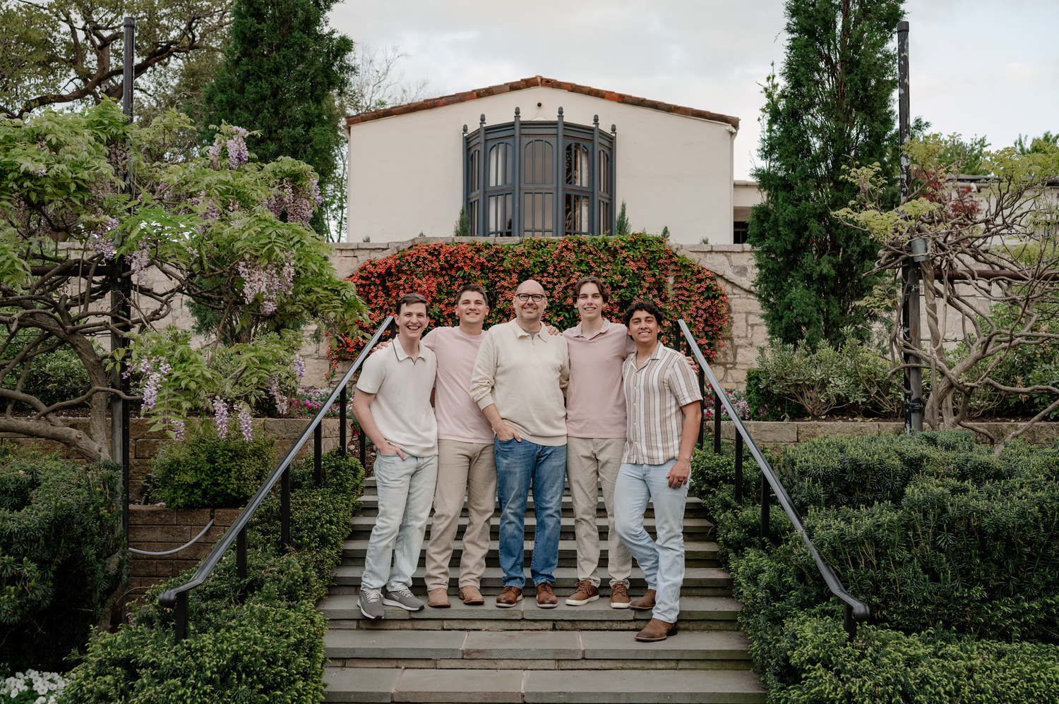 Group of men in a family standing on a staircase surrounded by greenery in front of a Spanish style house for their extended family session