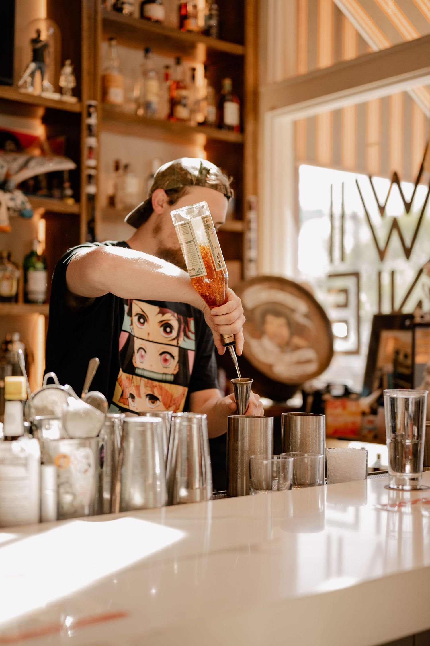 Bartender in casual attire pours a cocktail at bar counter with bottles displayed on shelves.