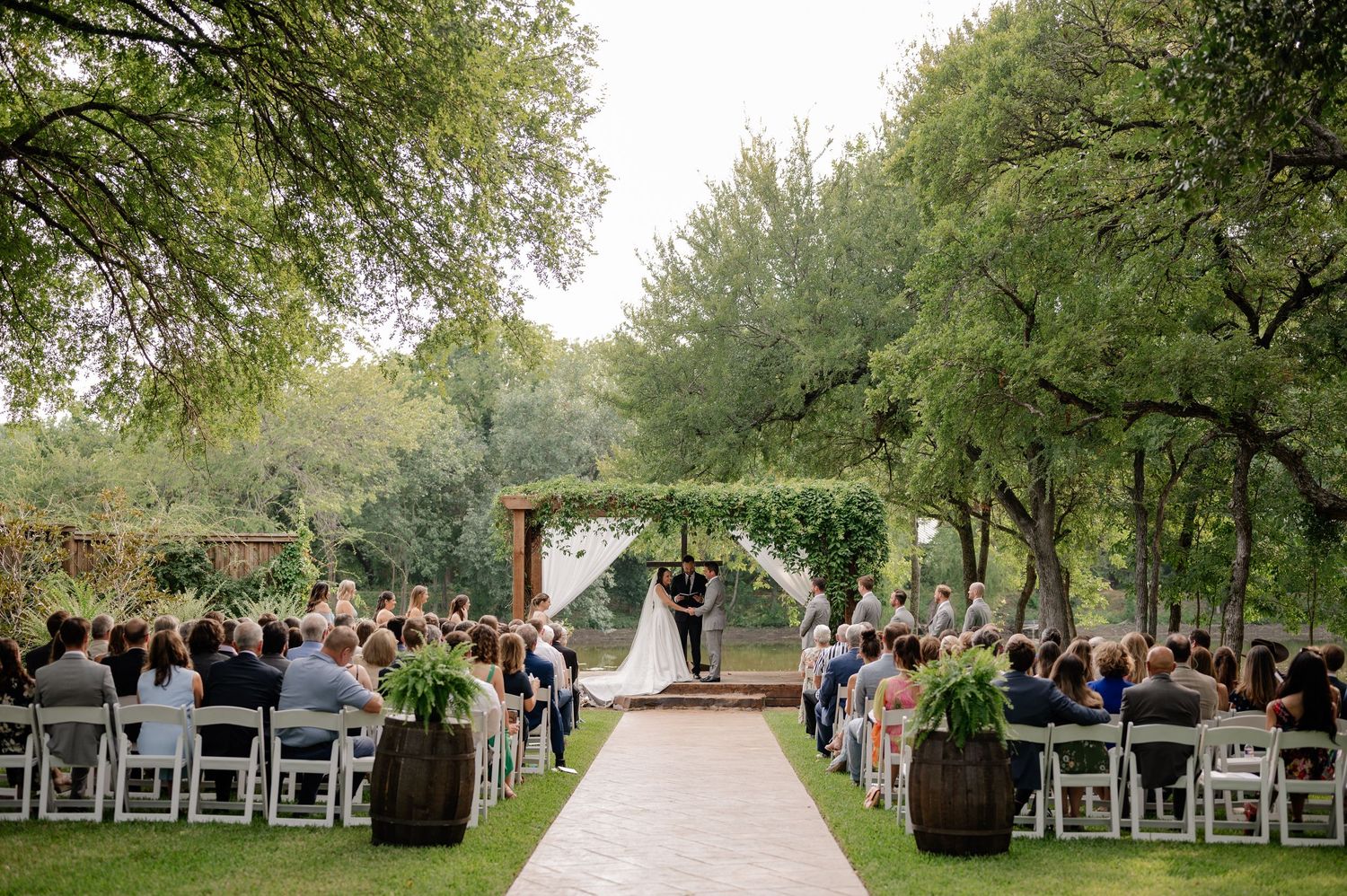 Dramatic wide shot bride and groom sharing vows during outdoor Spring wedding ceremony surrounded by greenery at venue in Plano, Texas