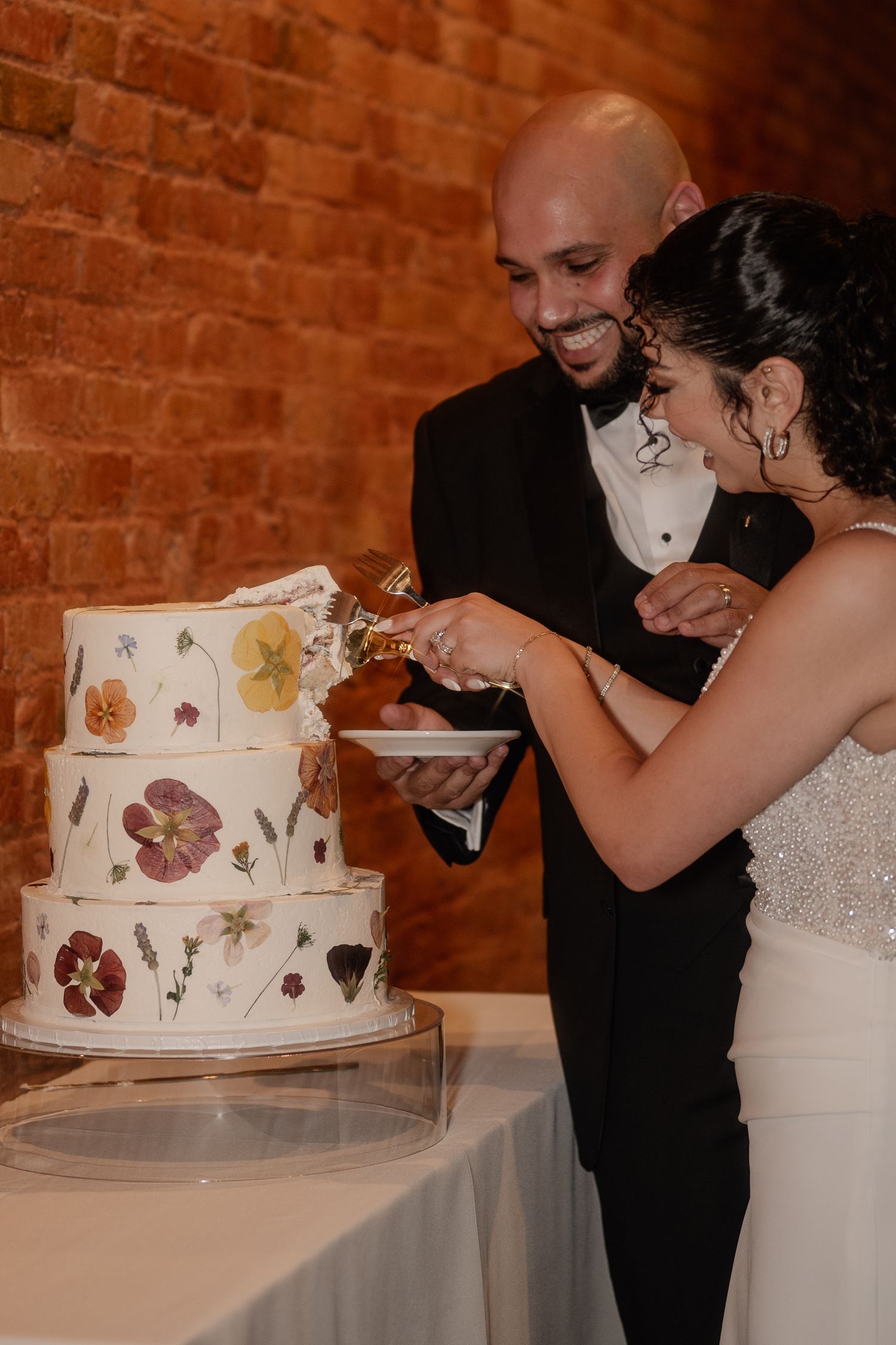 Bride and groom cutting into a beautiful floral cake during a lively reception in a moody brick walled wedding venue