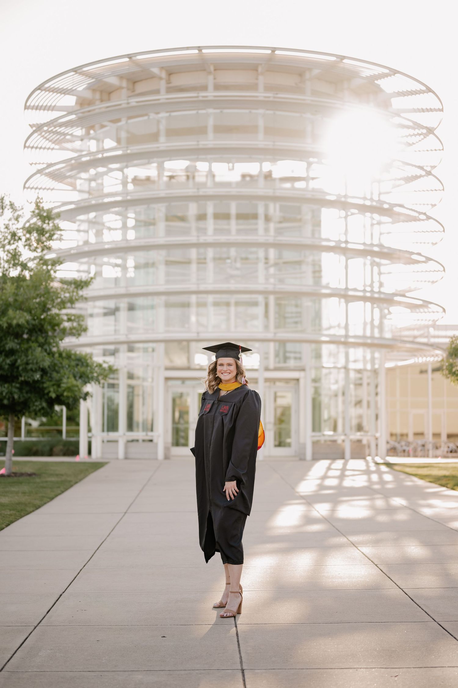 College senior in cap & gown posing in front of a white architectural building on the UT Dallas campus for a summer senior photo session
