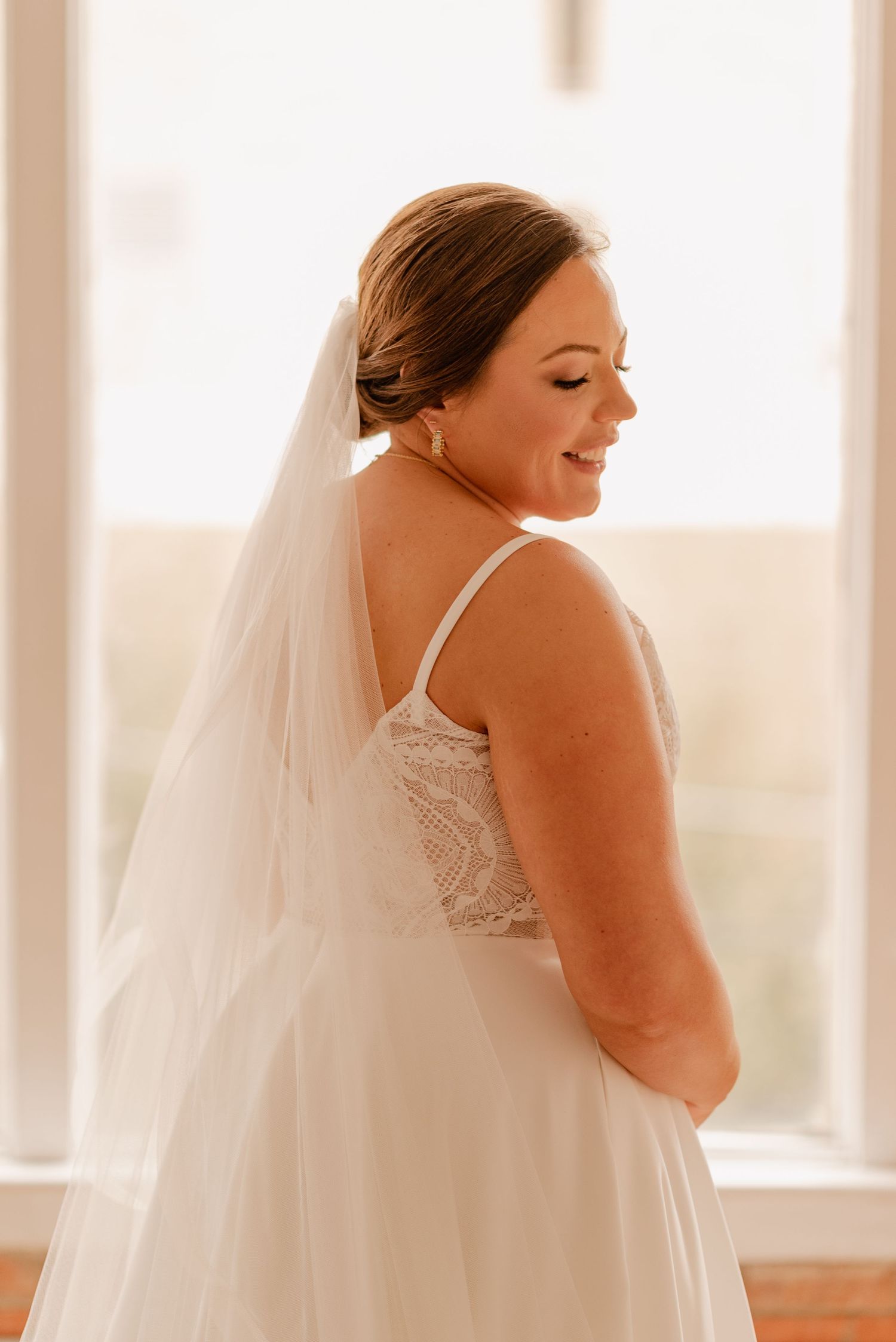 Side profile of bride wearing delicate veil and lace-detailed wedding dress in natural window light.