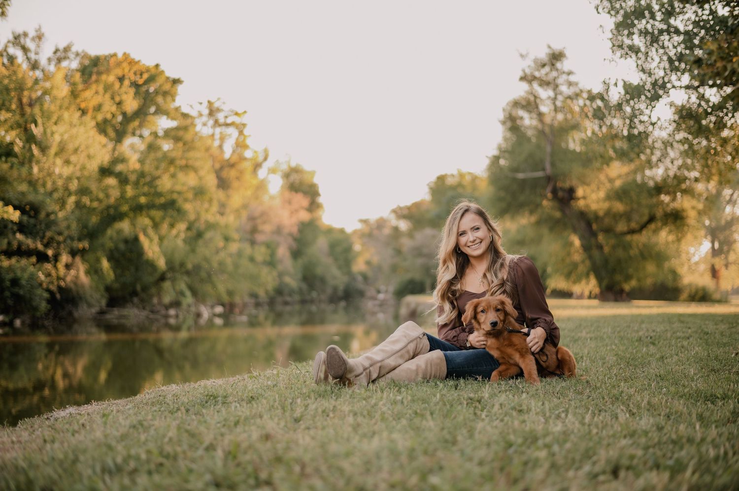 Woman with her puppy sitting in front of a tree lined lake in Dallas, TX for a fall portrait session