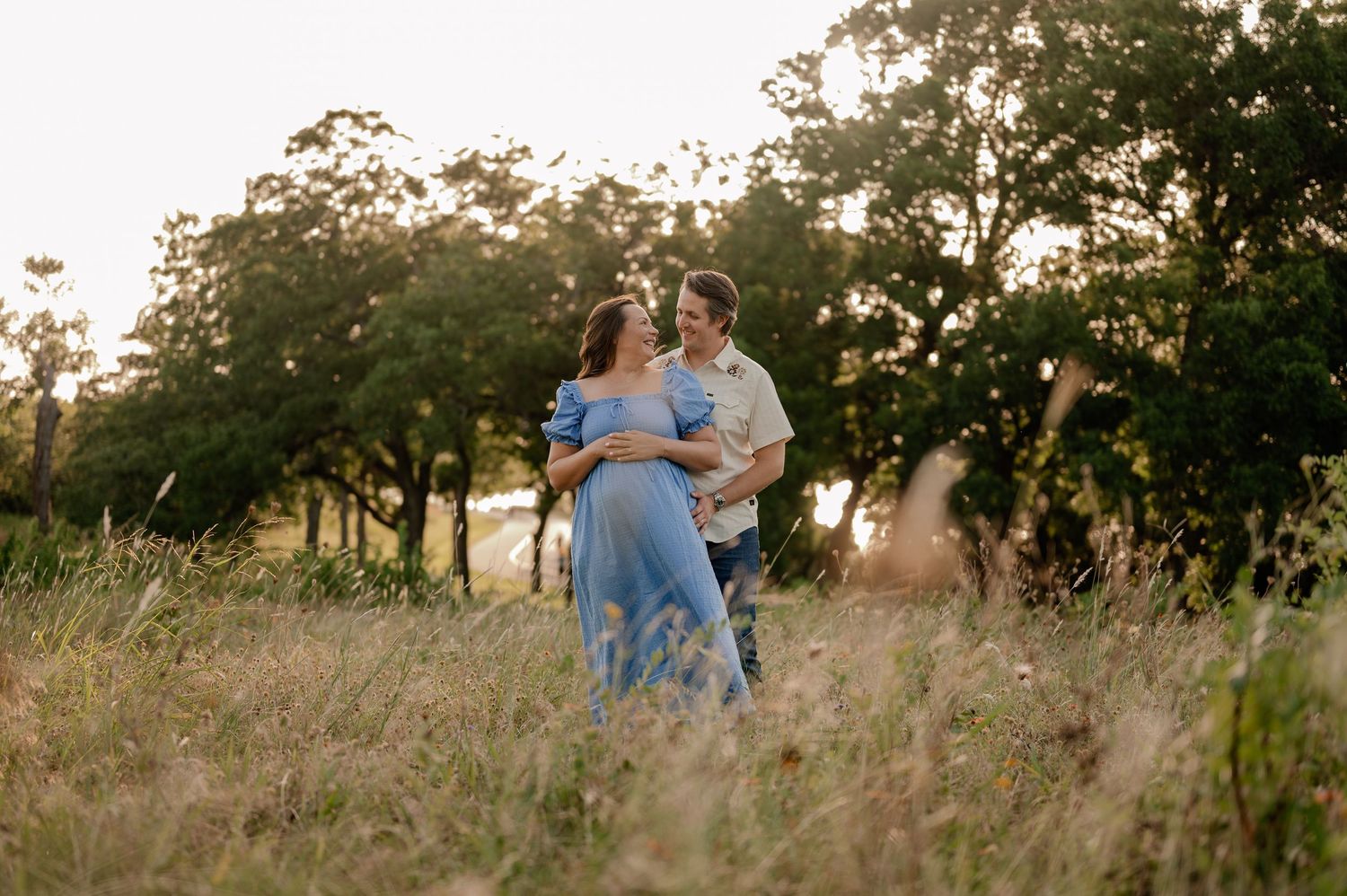 Candid photo of a husband holding his wife while she holds her pregnant belly at a summer maternity session overlooking a lake in Dallas, TX