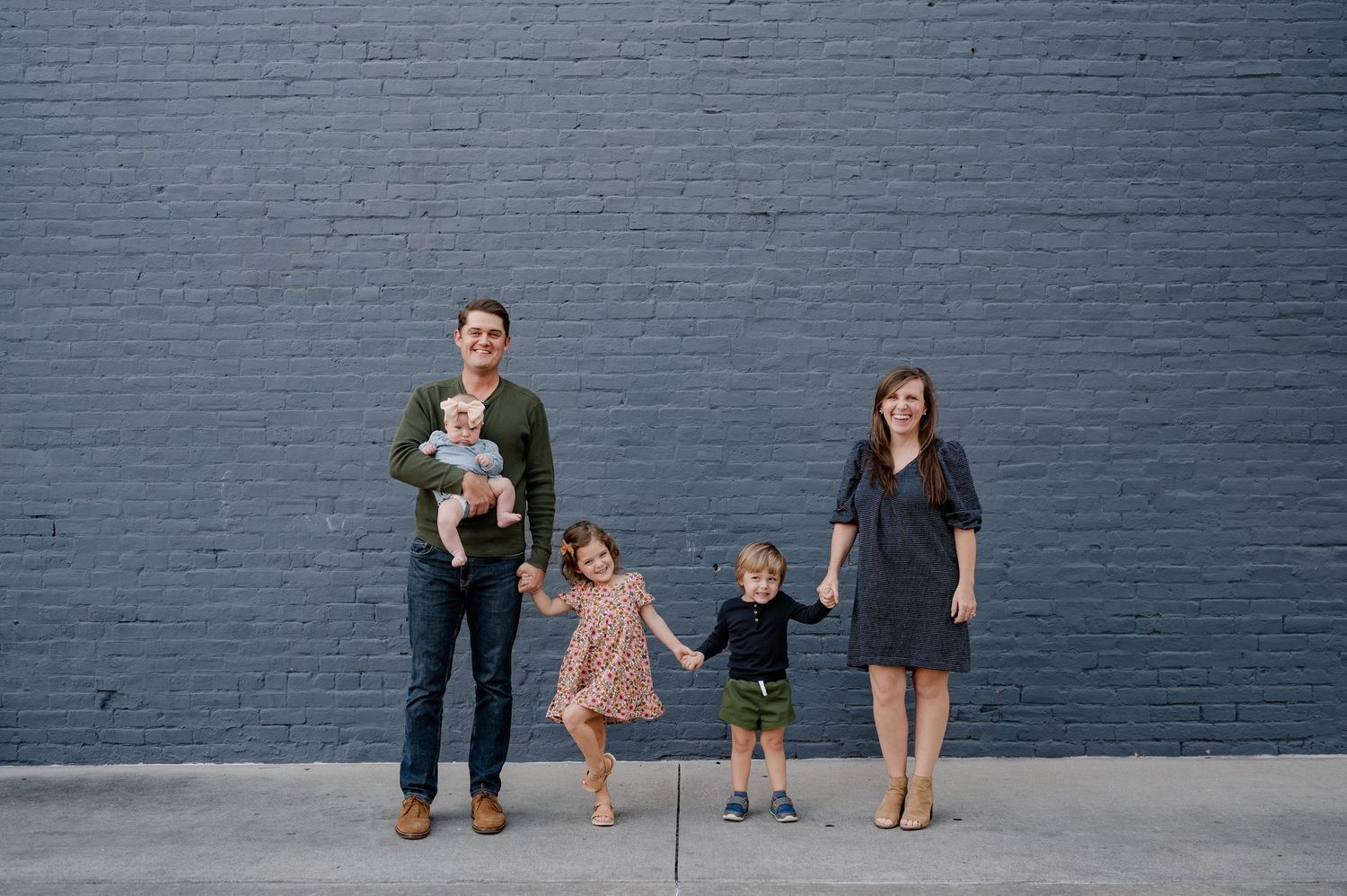 Family of 5 holding hands and posing in front of a blue painted brick wall for fall family photos in downtown Rockwall