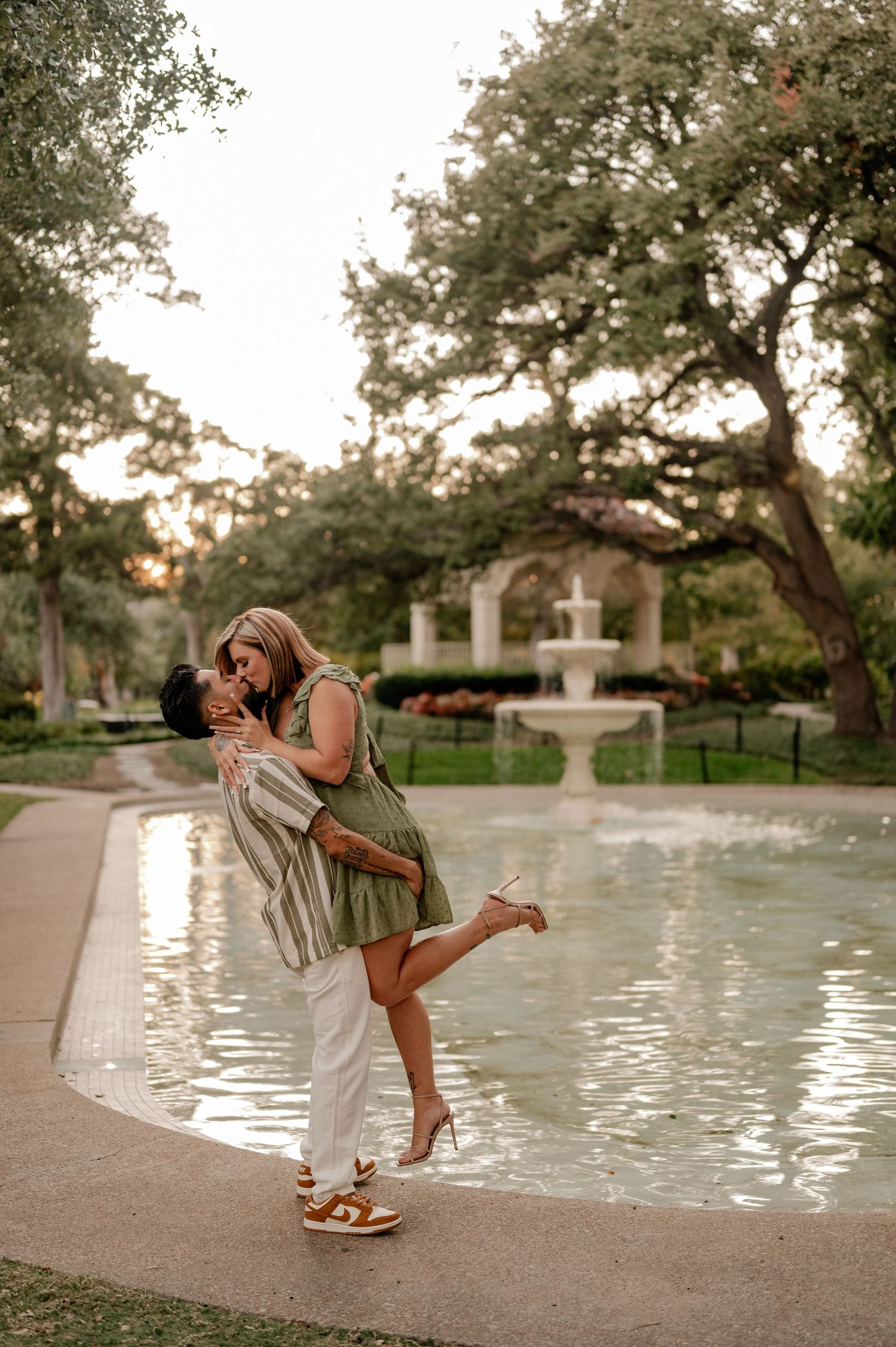 Couple sharing a romantic kiss by a fountain in a lush garden, woman in green dress lifted by partner at Flippen Park in Dallas, Texas.