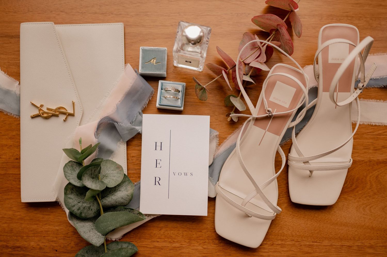 Wedding flat lay with white strappy sandals, YSL logo, and eucalyptus on wooden surface.