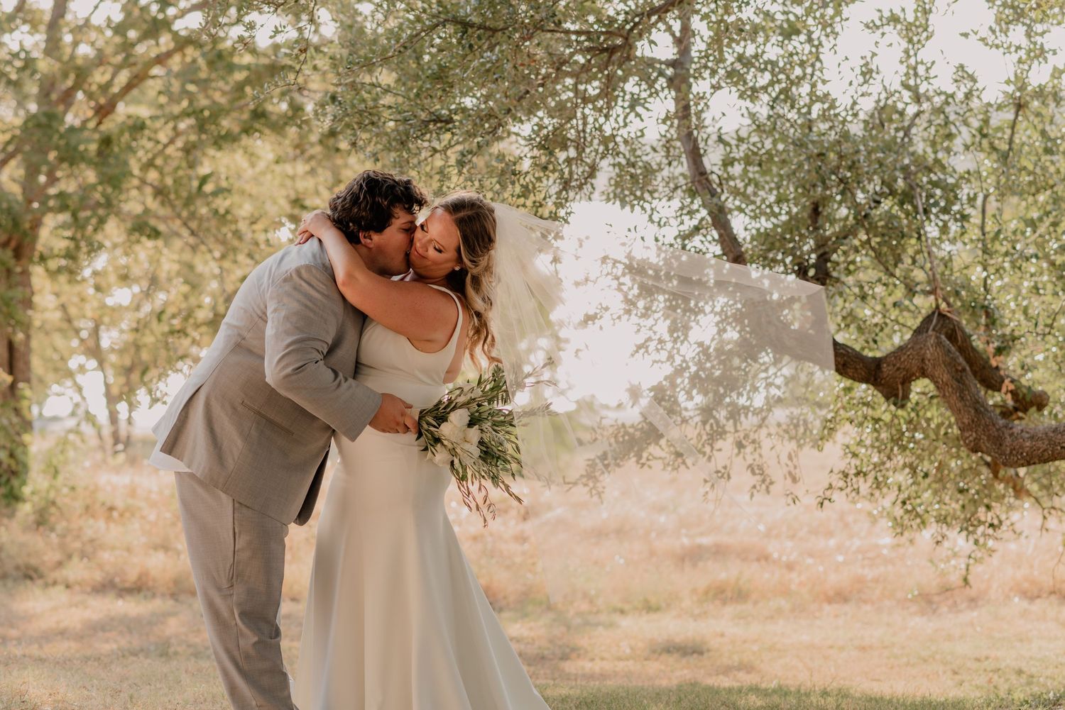 A couple in wedding attire share an intimate moment under a sunlit tree in an outdoor setting.