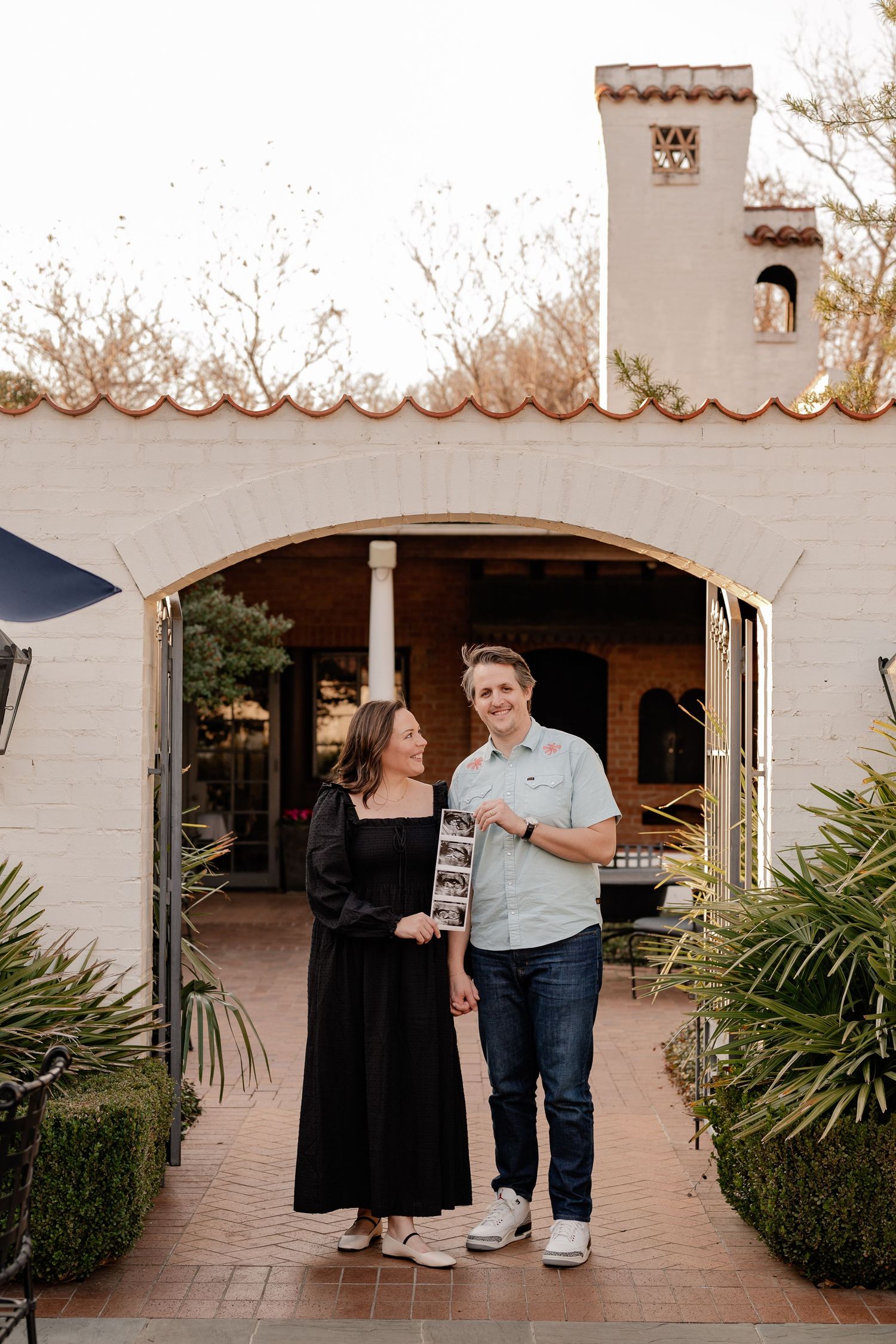 Couple posing with sonogram of their new baby under a Spanish style archway for a Spring baby announcement at the Dallas Arboretum