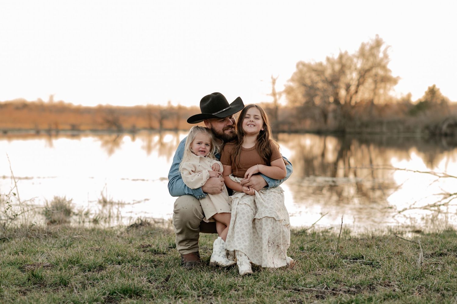 Father hugging his daughters in front of a large pond on their Rockwall Texas farm for a fall at-home family photo session at golden hour
