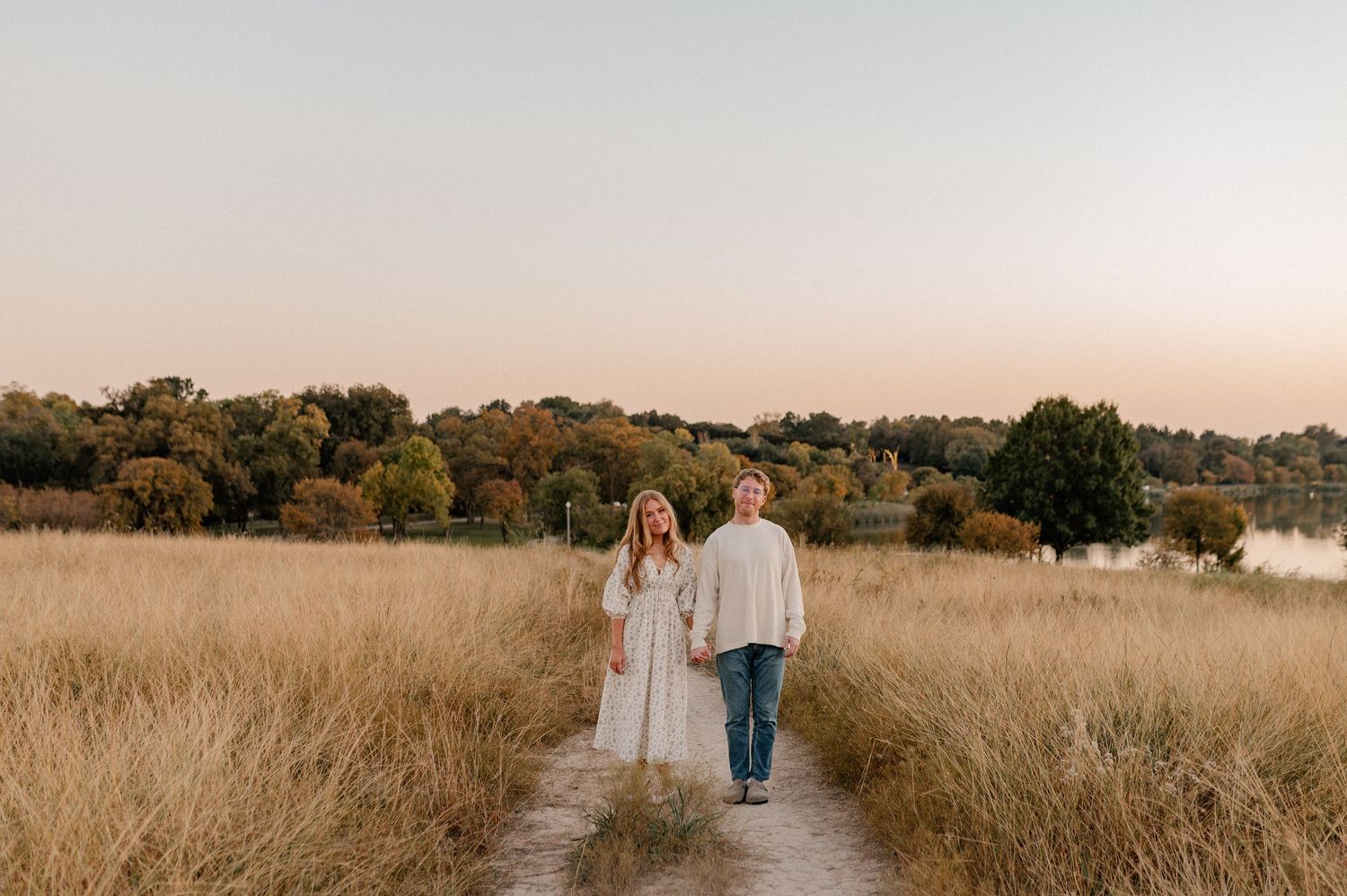 Posed photo of couple handing hands in the tall grass of a field overlooking White Rock Lake during golden hour in East Dallas, Texas