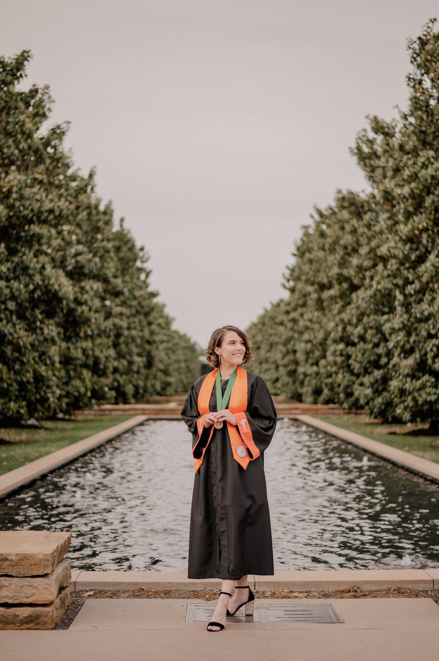 College senior in grad gown posing in front of a treelined  on the UT Dallas campus for a summer senior photo session