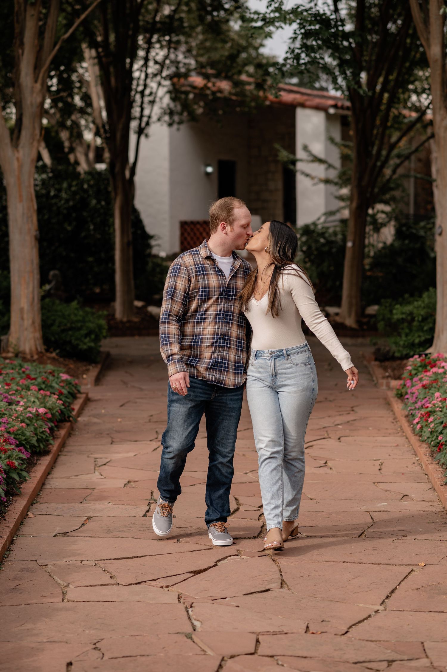 Couple sharing a kiss while walking on a stone pathway surrounded by trees and flowers in a garden at the Dallas Arboretum