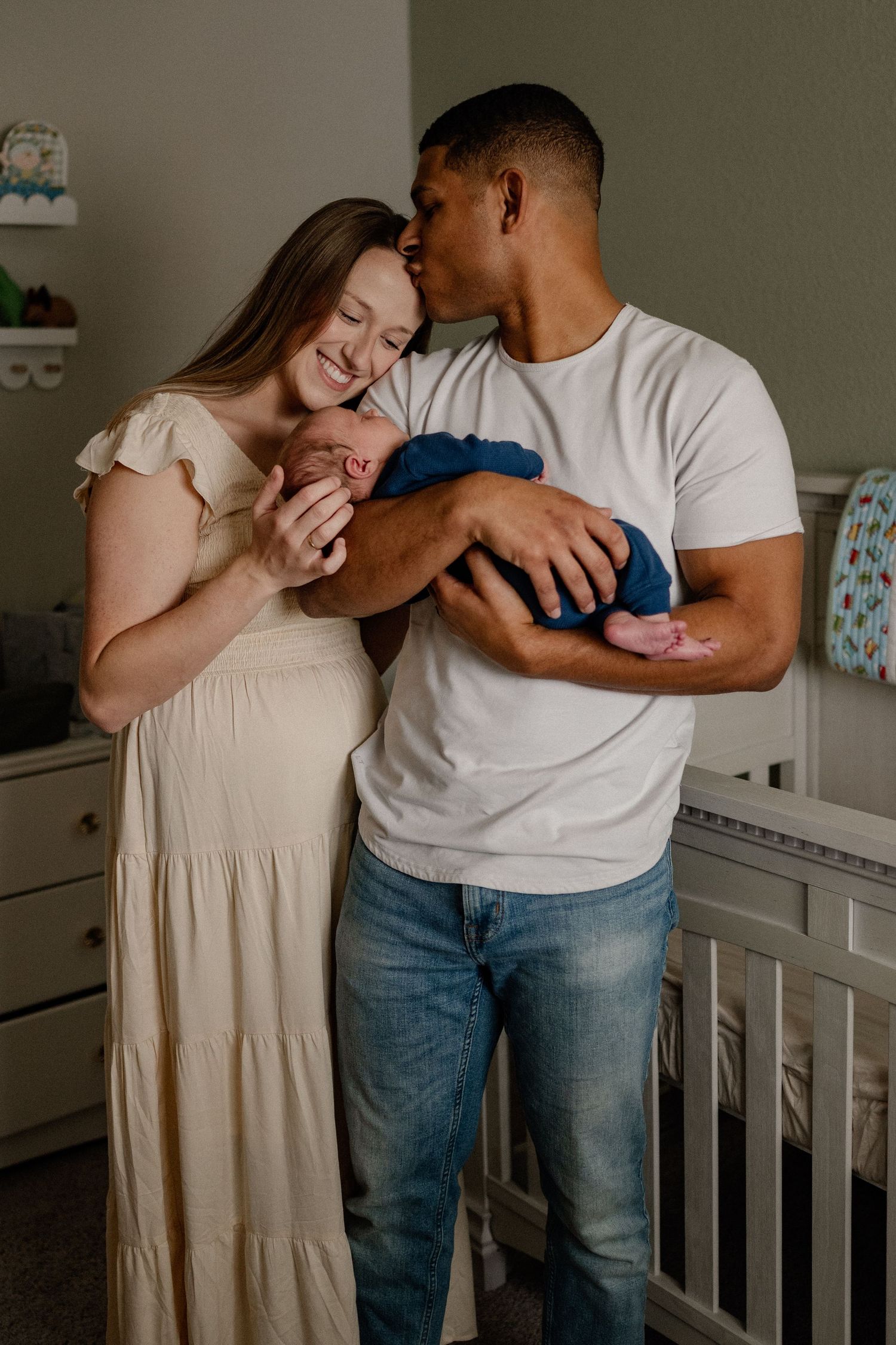 Husband kissing wife's forehead while she's staring at newborn in her husband's arms in their nursery for an at-home newborn photo session