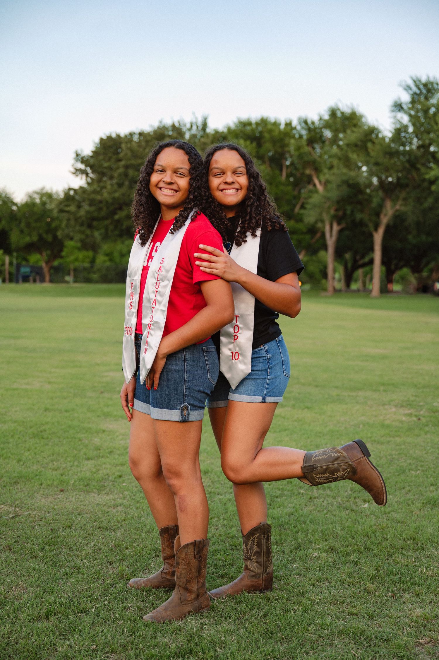 Senior twins in their graduation awards standing in a large tree-filled park in Richardson, Texas for a senior photo session