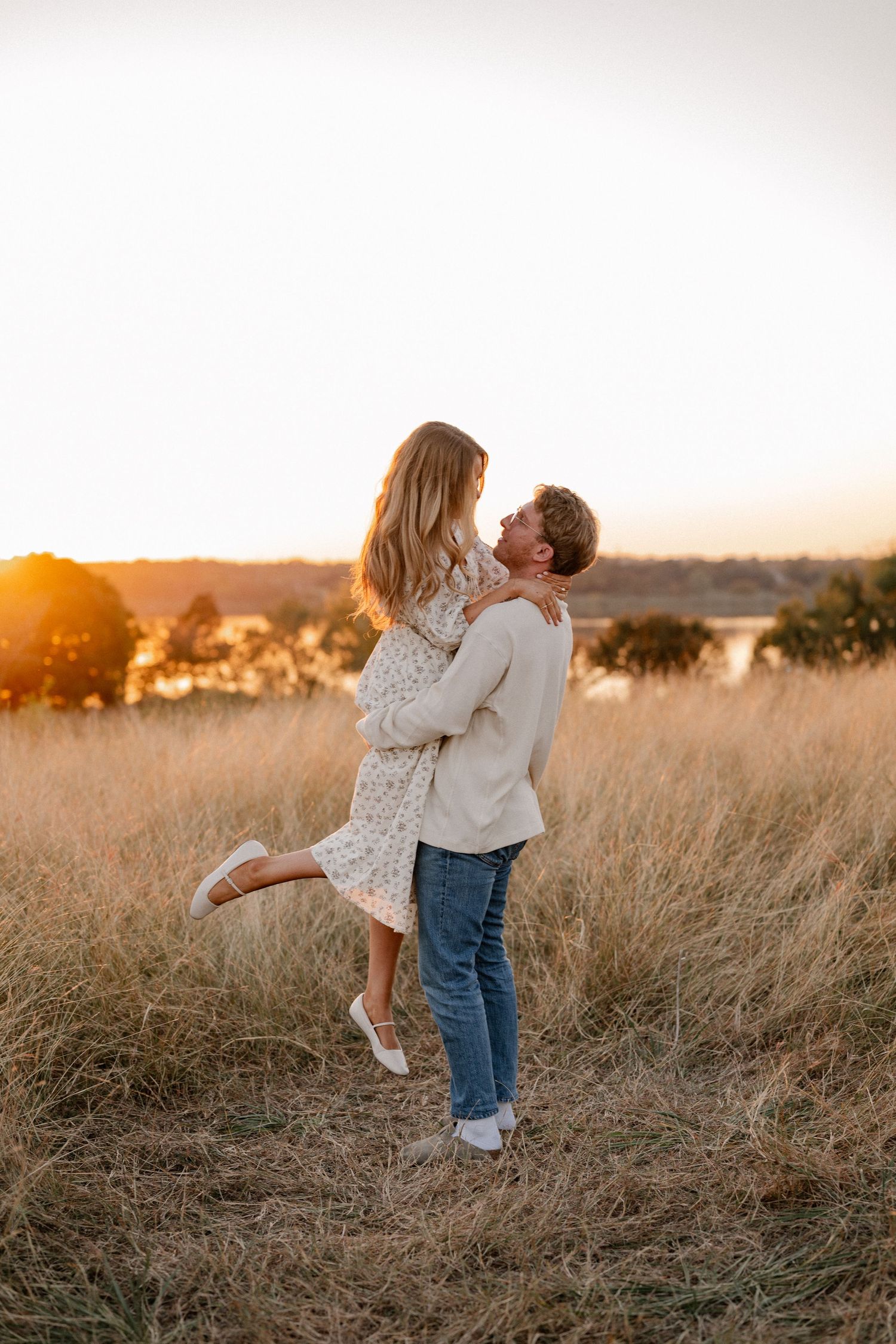 Romantic photo of a man picking up and spinning his fiance at a large tall grass field overlooking the lake in East Dallas at golden hour