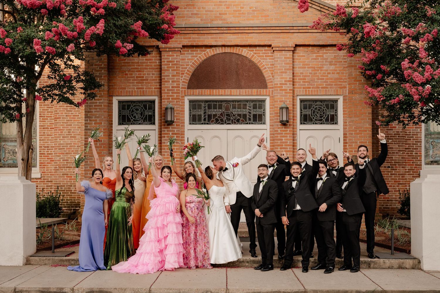 Large wedding party celebrating while the bride and groom kiss outside of the large red bricked catholic church in Natchitoches, Louisiana