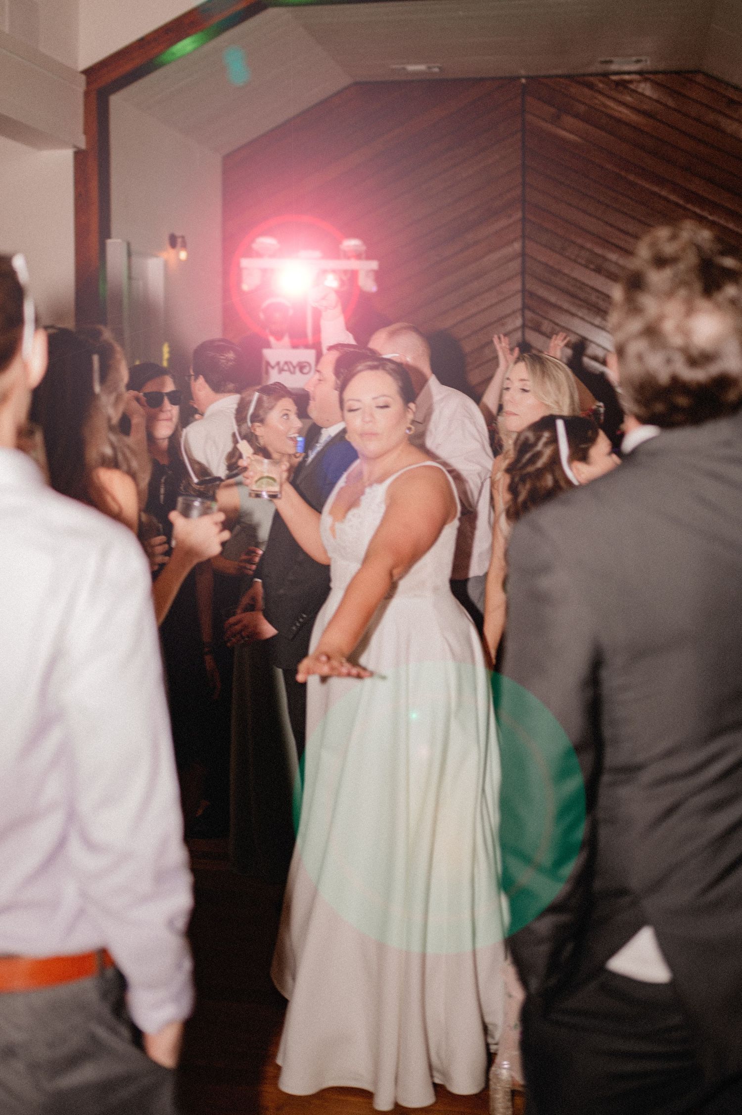 Dance floor scene of bride in sunglasses at wedding reception with colorful lighting effects.