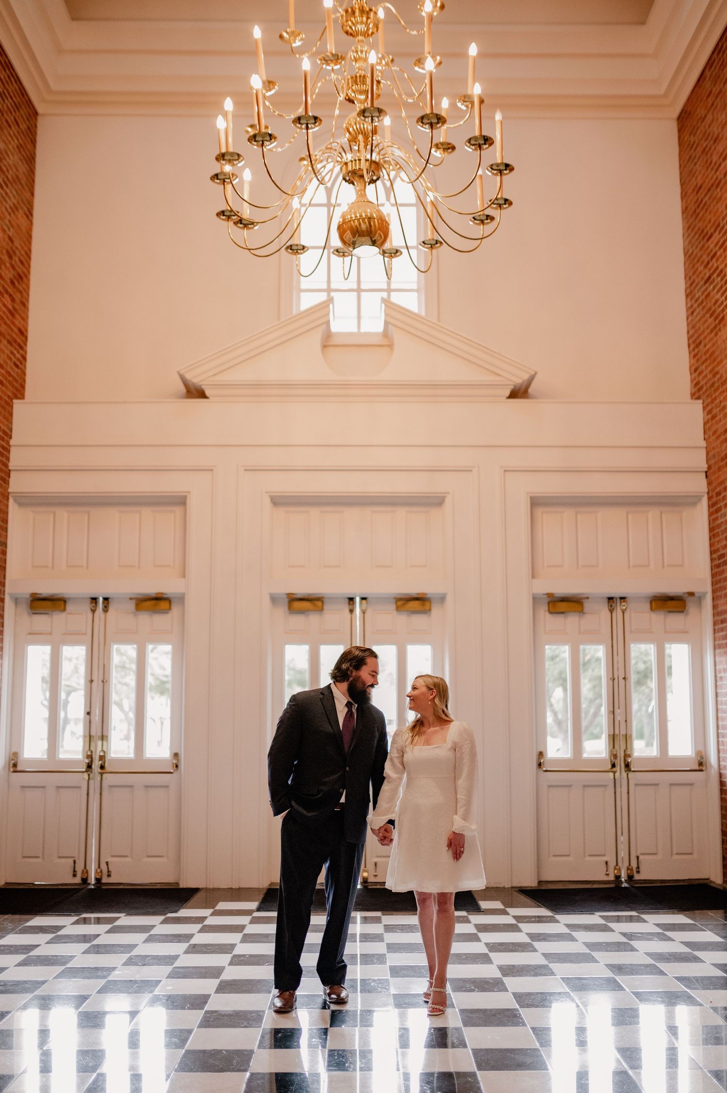Elegant chandelier hangs in a grand hall with black and white checkered floors and white paneled walls.