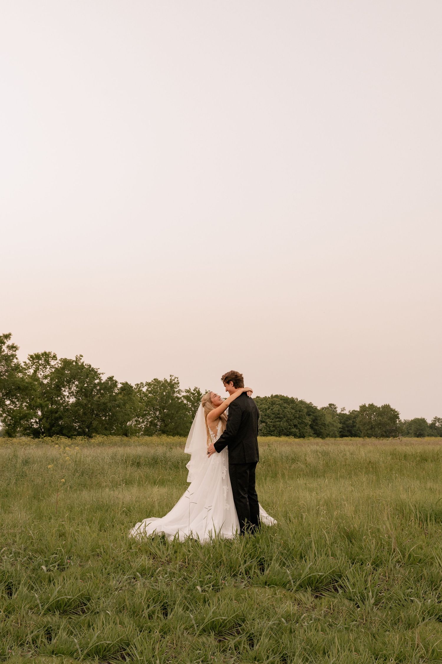 Candid documentary style photo of bride and groom embracing in wide green field during couple's wedding portraits at venue in Rockwall, TX