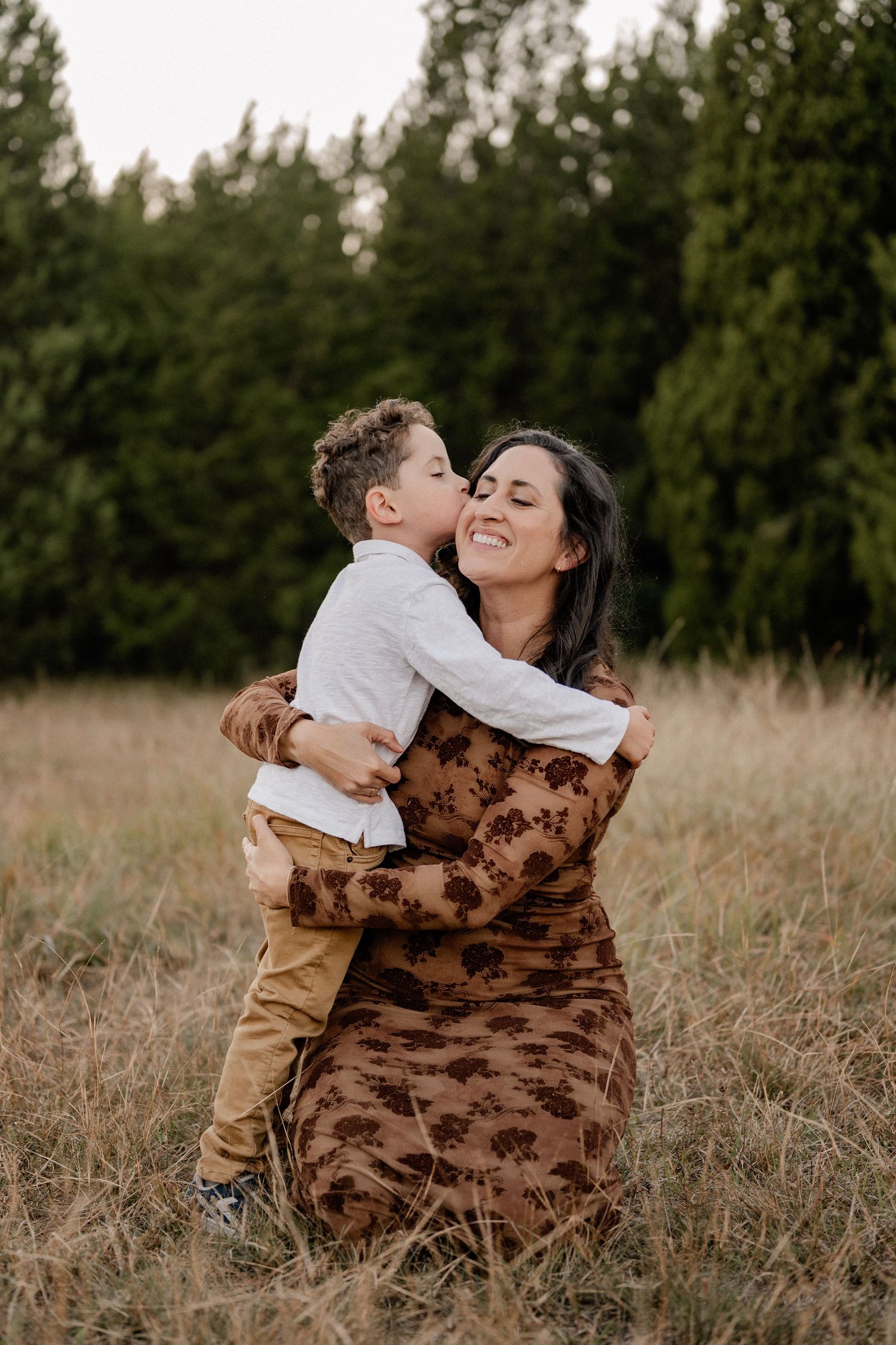 Son kissing on his mom in a large field lined with trees at an outdoor family session for fall photos in Sunnyvale, Texas