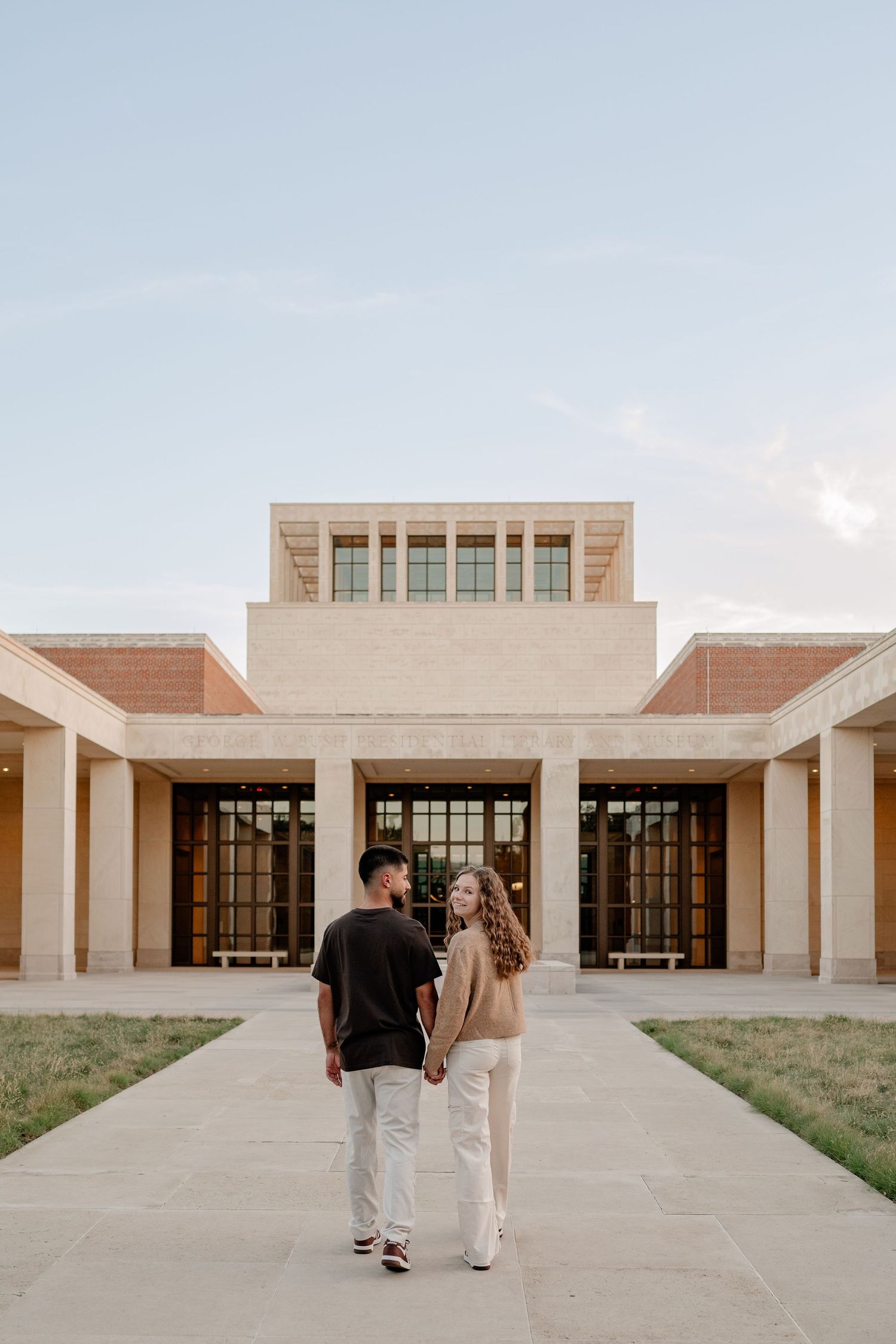 Couple holding hands walking toward large architectural building on the SMU campus in Dallas Texas for a fall engagement photo session
