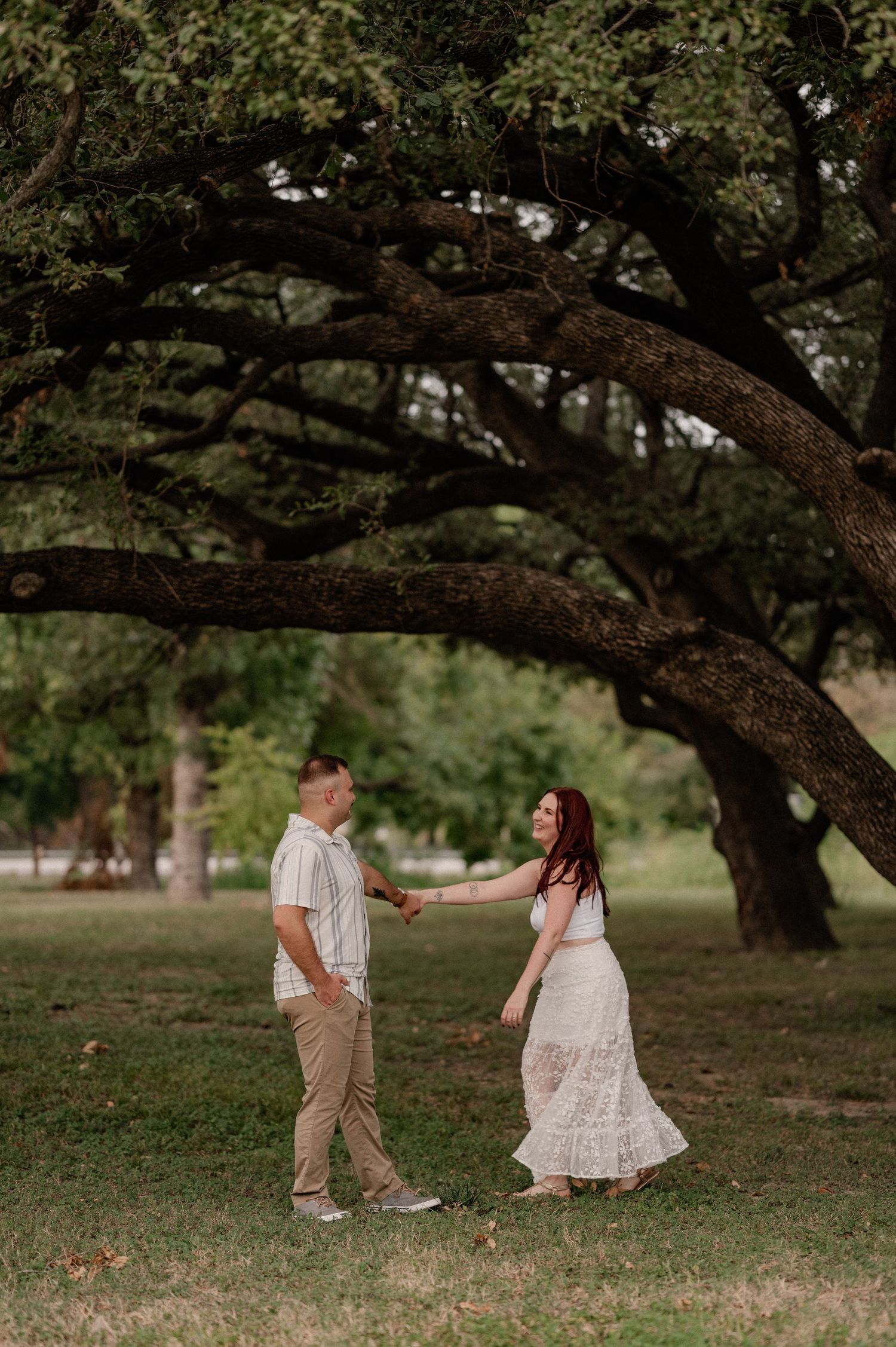 Couple candidly dancing and spinning under a large shady tree during their summer engagement session in a wide Dallas field