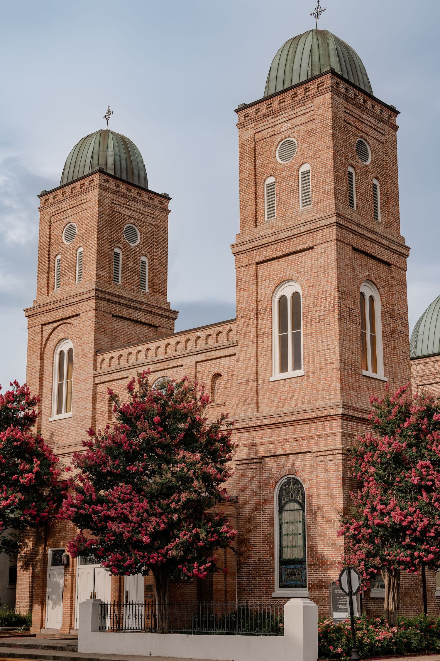 Historic red brick church with twin copper domed towers and crosses, framed by pink crepe myrtle trees in Natchitoches, Louisiana